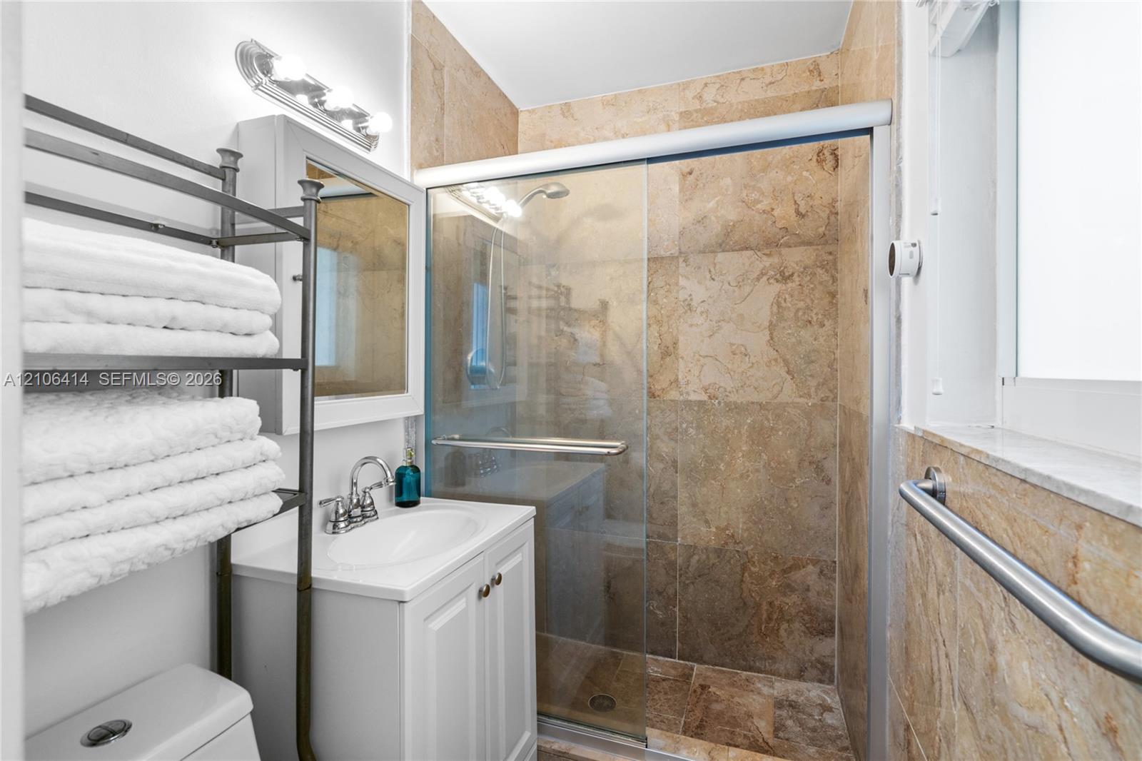 Bathroom featuring a white vanity cabinet with an integrated sink, a glass-enclosed shower with full-height stone-finish tile surround, and an attached wall-mounted metal grab bar