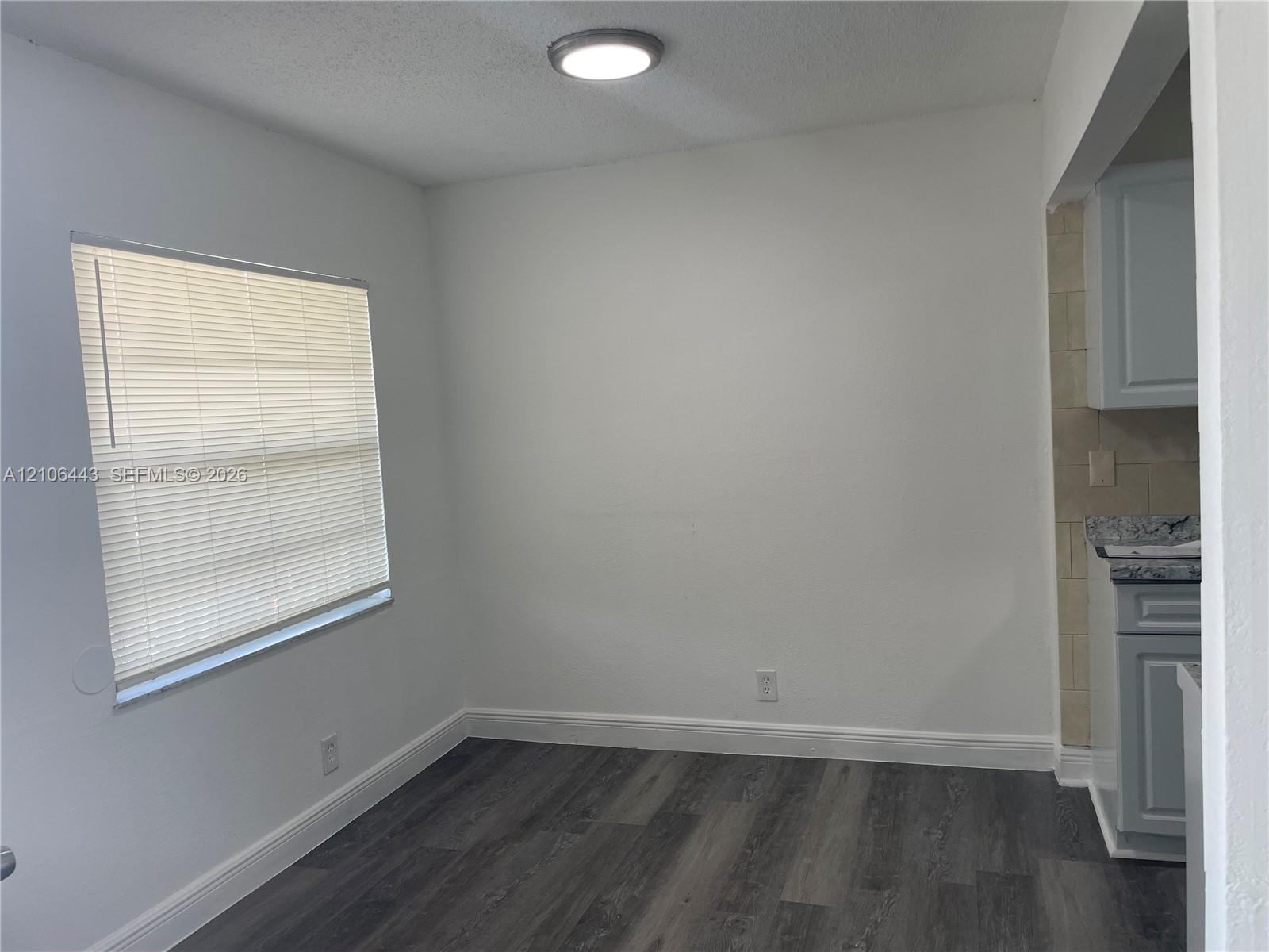Room featuring wood-finish flooring, a window with installed blinds, and a flush-mount ceiling light fixture, accented by cabinetry with stone-pattern countertops