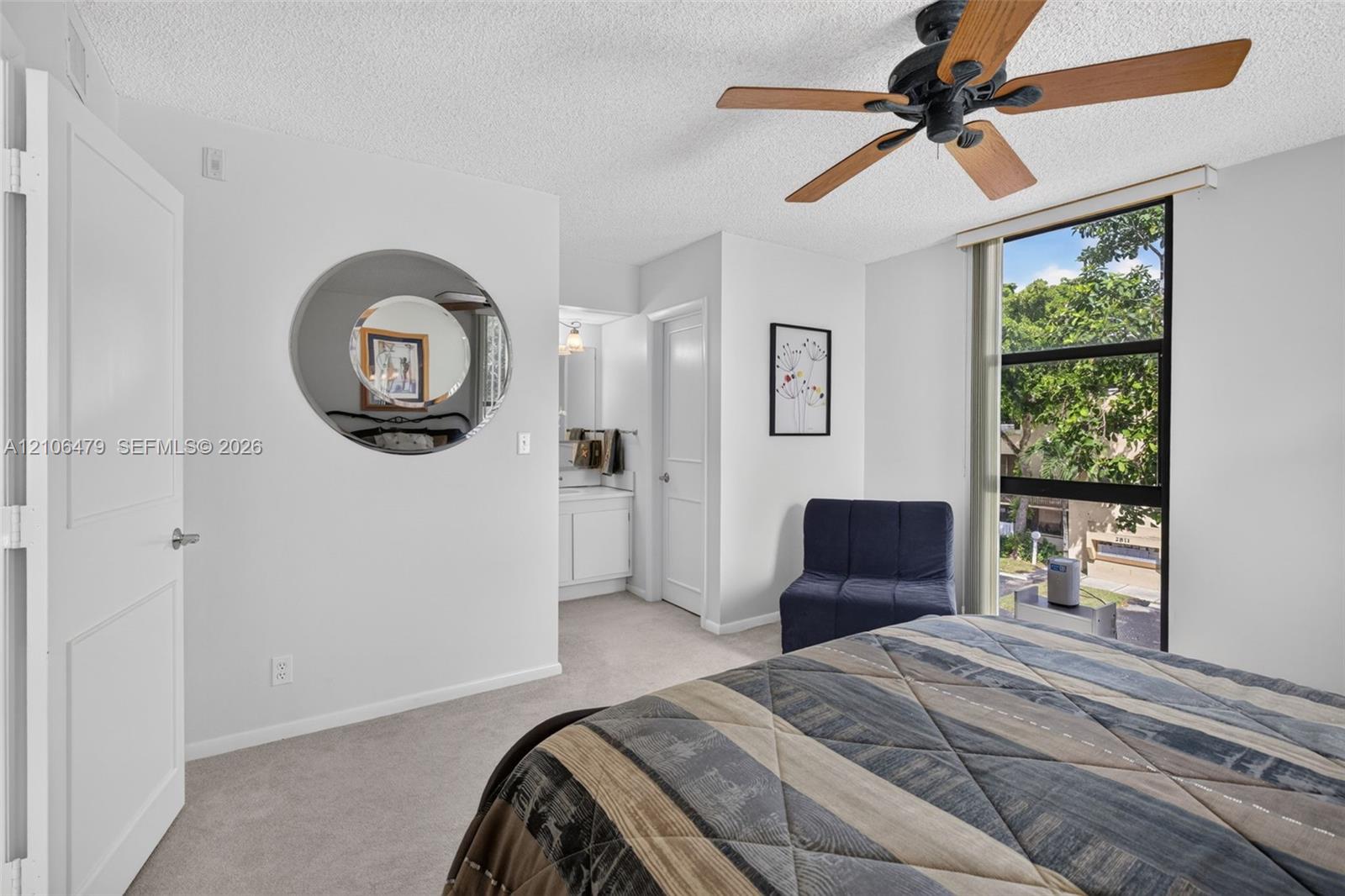Neutral wall-to-wall carpeting, multi-panel interior door, ceiling fan with wood-finish blades, and large fixed-pane window with black framing
