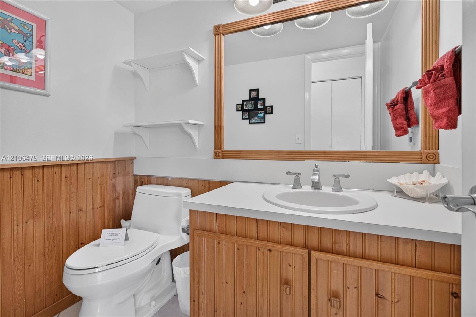 Vanity featuring a solid surface countertop, integrated oval basin, and wood-finish cabinetry with wainscoting, accented by a large wood-framed mirror and wall-mounted shelving