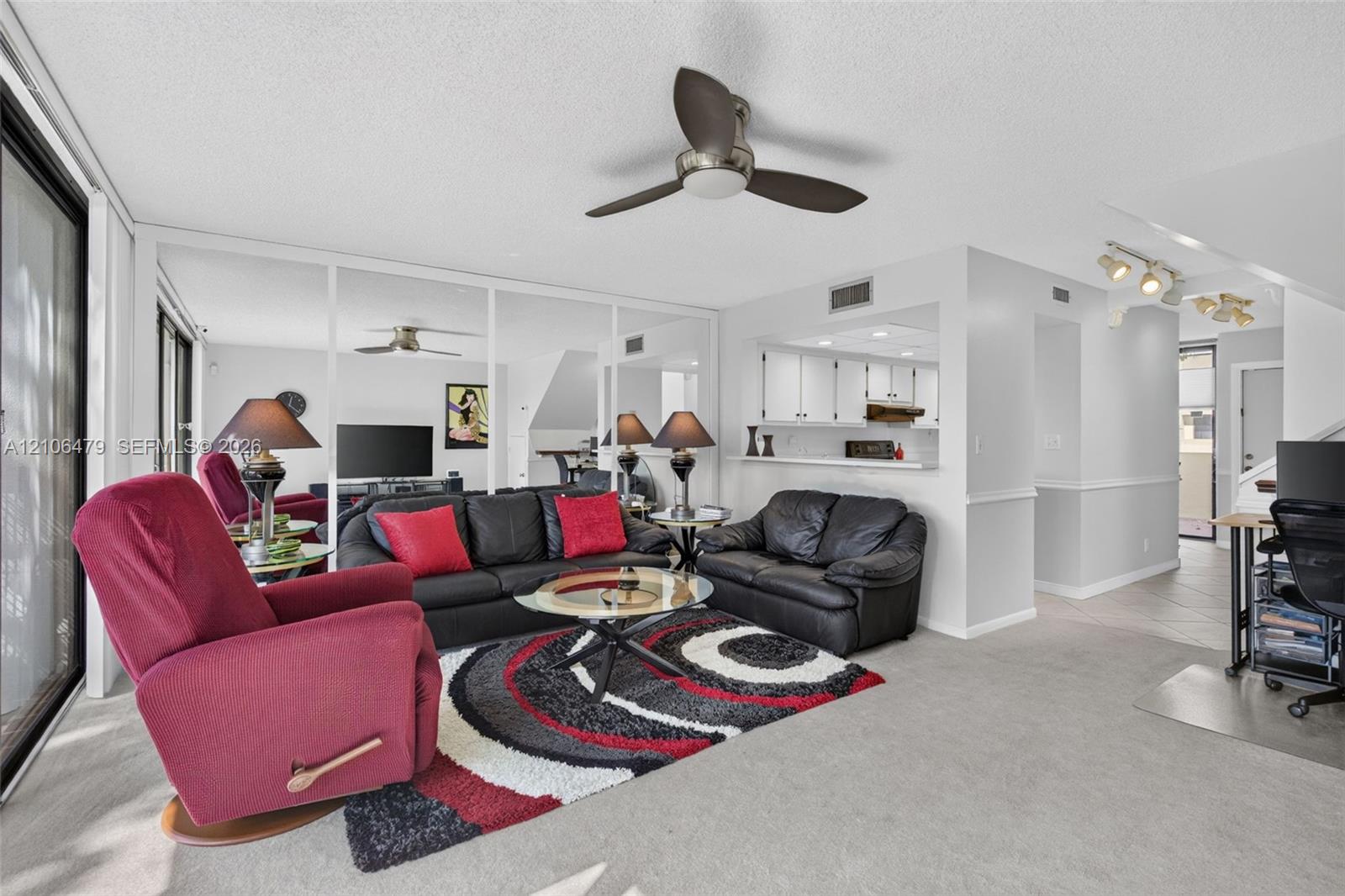 Living area featuring a ceiling fan, wall-to-wall mirrored panels, carpeted flooring, and a pass-through opening to the kitchen