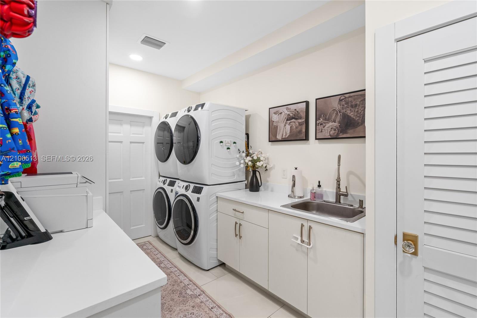 Laundry room featuring stacked washer and dryer units, solid-surface countertops, an undermount stainless steel sink with a high-arch faucet, custom flat-panel cabinetry, and louvered door access