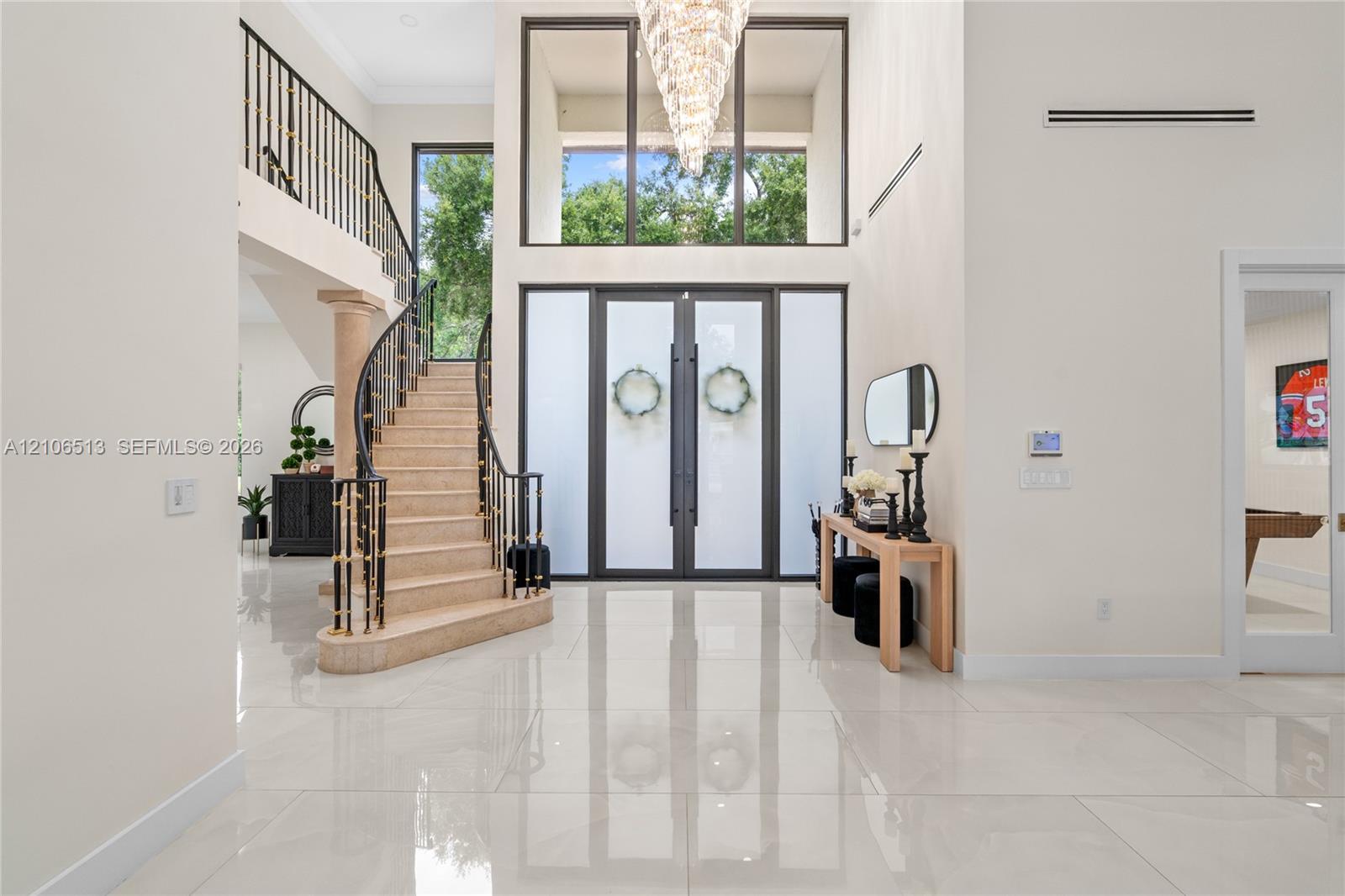 Grand foyer featuring polished large-format tile flooring, double entry doors with frosted glass panels, an expansive multi-story window, a tiered crystal chandelier, and a curved staircase with metal railings