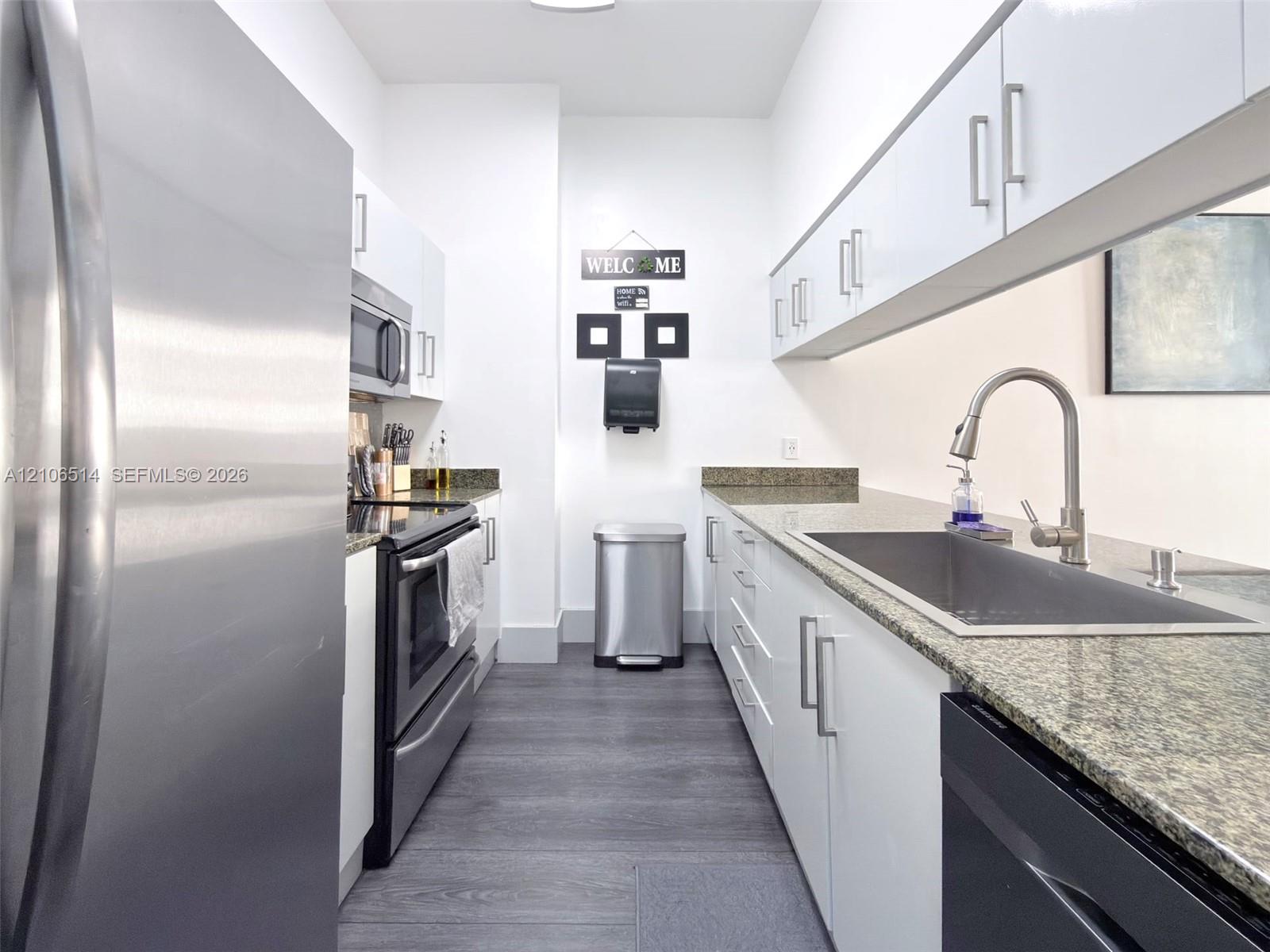 Galley-style kitchen featuring high-gloss white cabinetry, stone countertops, stainless steel appliances, and wood-finish flooring