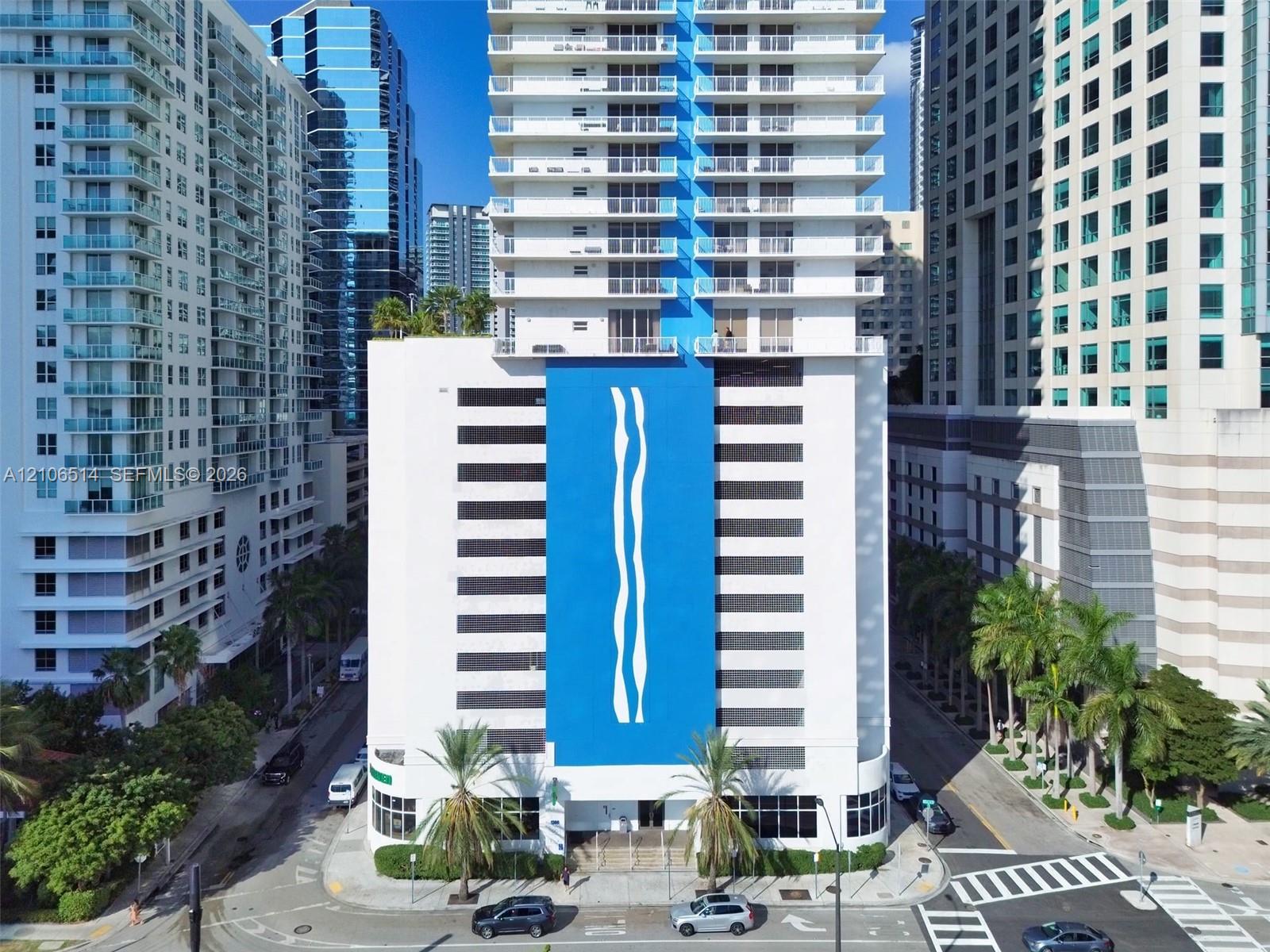 High-rise residential exterior featuring a blue vertical accent wall, multiple private balconies, ground-level landscaping with palm trees, and floor-to-ceiling windows