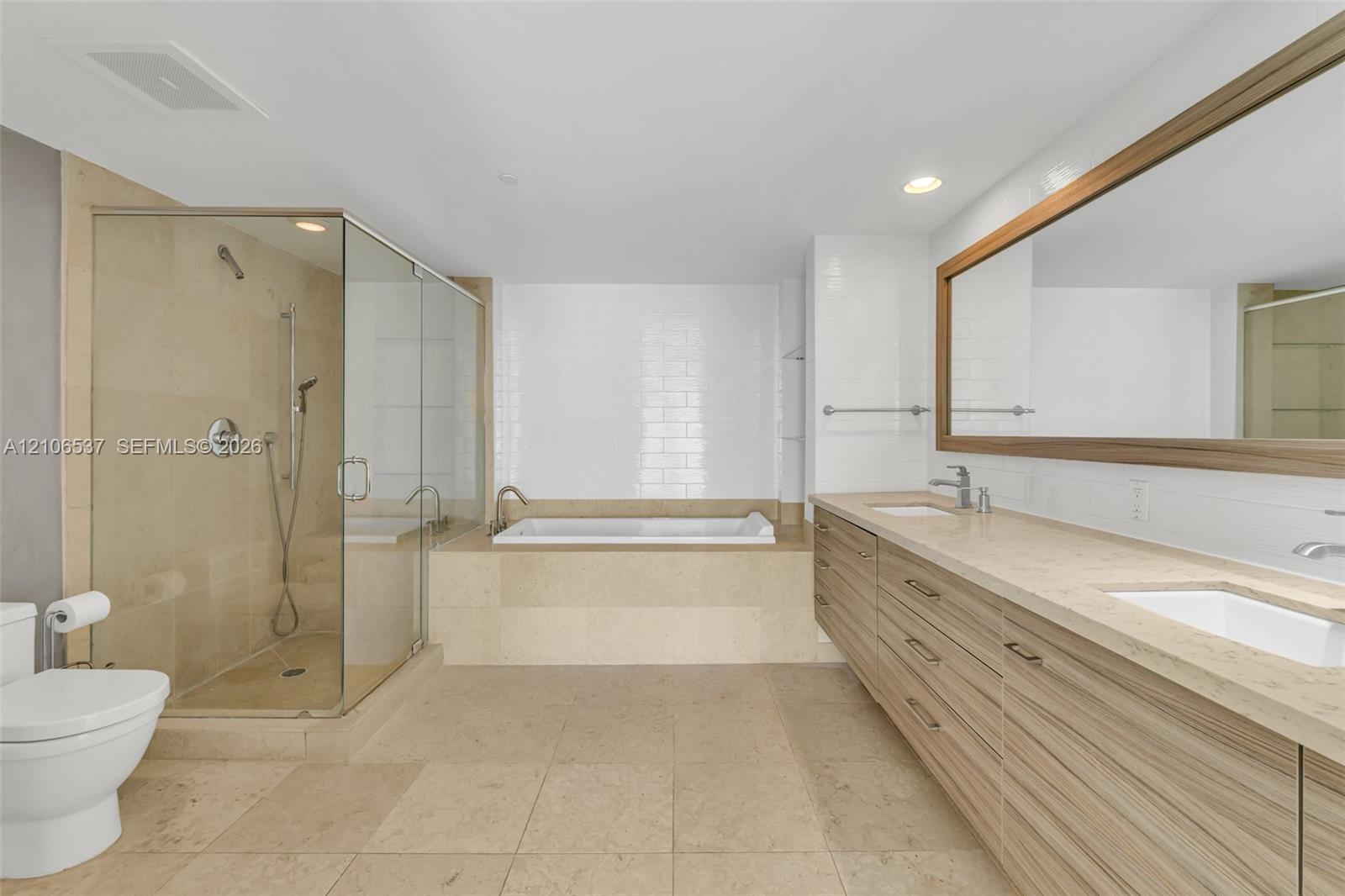 Bathroom featuring dual-sink vanity with wood-finish cabinetry, stone-style countertop, glass-enclosed shower, and a built-in soaking tub with white tile surround