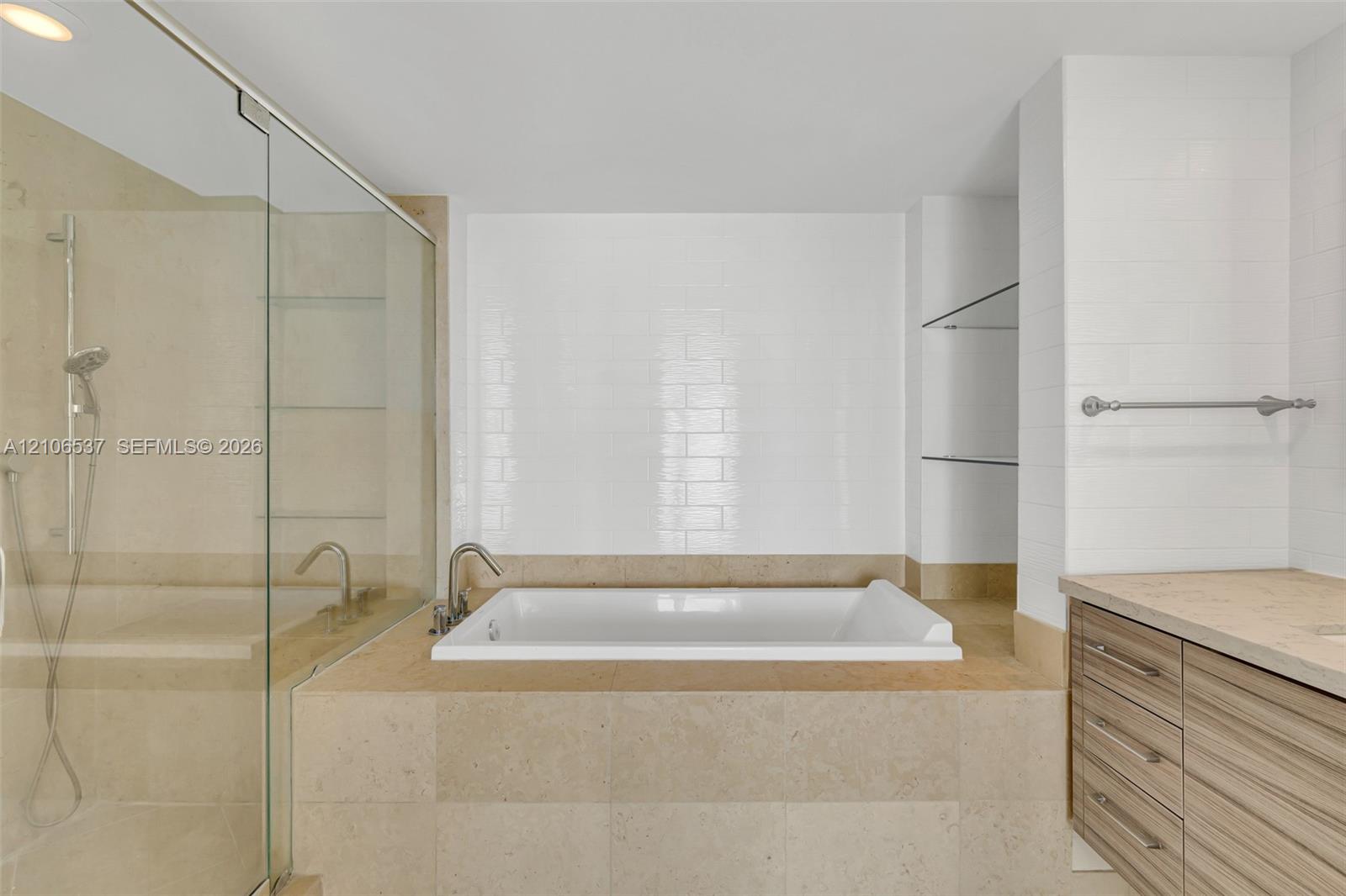 Soaking tub featuring a tiled surround, glass-enclosed shower, wall-mounted faucet fixtures, wood-finish cabinetry, and built-in glass shelving