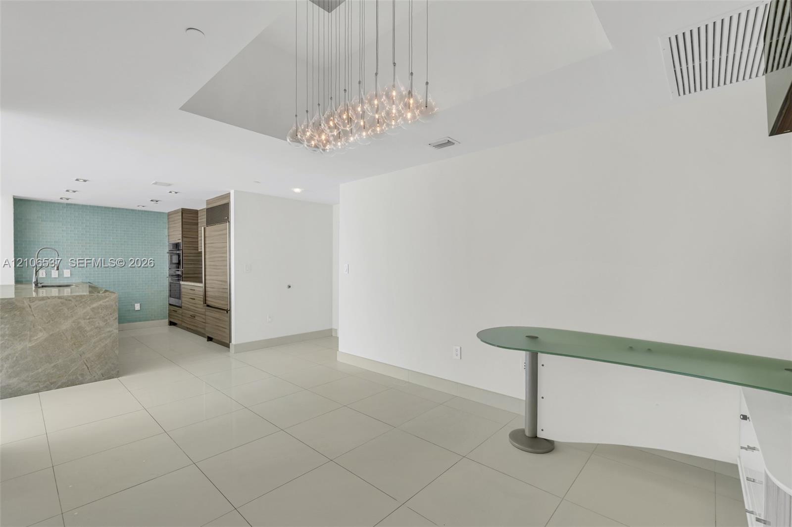 Large-format tile flooring, multi-pendant glass chandelier, built-in stone-surfaced kitchen island, wall-mounted curved glass console, and floor-to-ceiling wooden cabinetry with integrated appliances