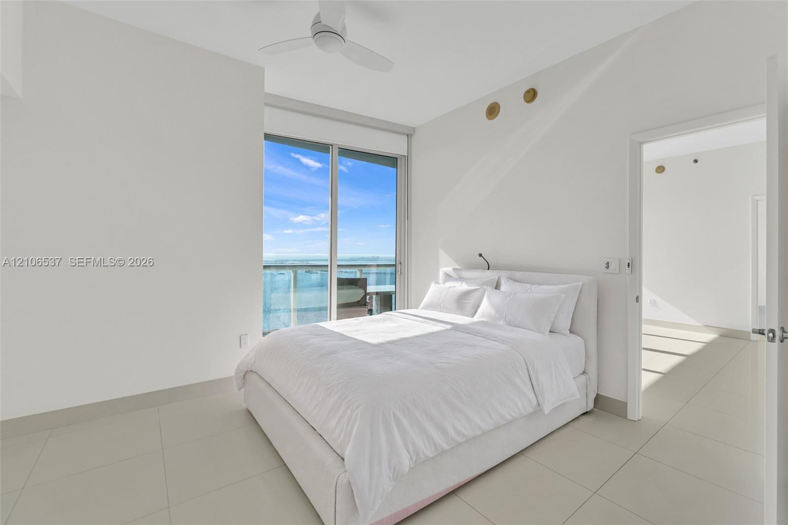 Large-format tile flooring, ceiling fan with integrated lighting, and floor-to-ceiling glass sliding doors leading to a private balcony with water scenery
