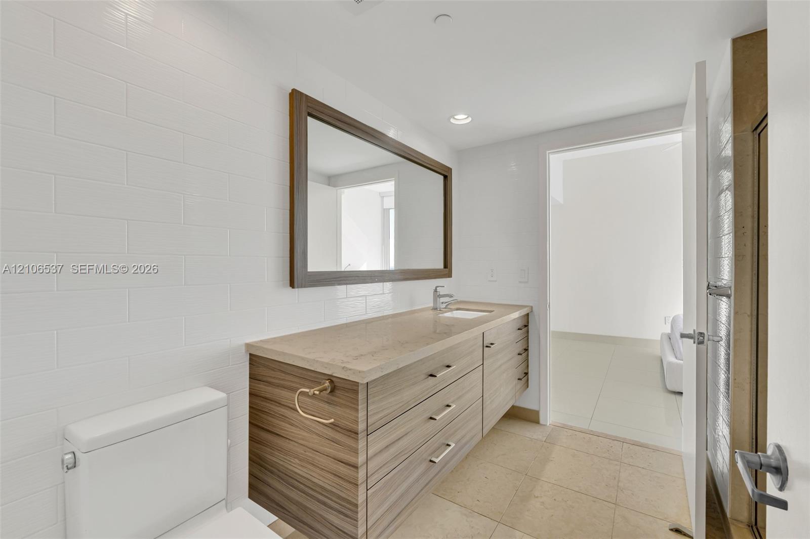 Floating vanity with wood-finish cabinetry, stone countertop, integrated sink, framed wall mirror, and subway-tiled wall
