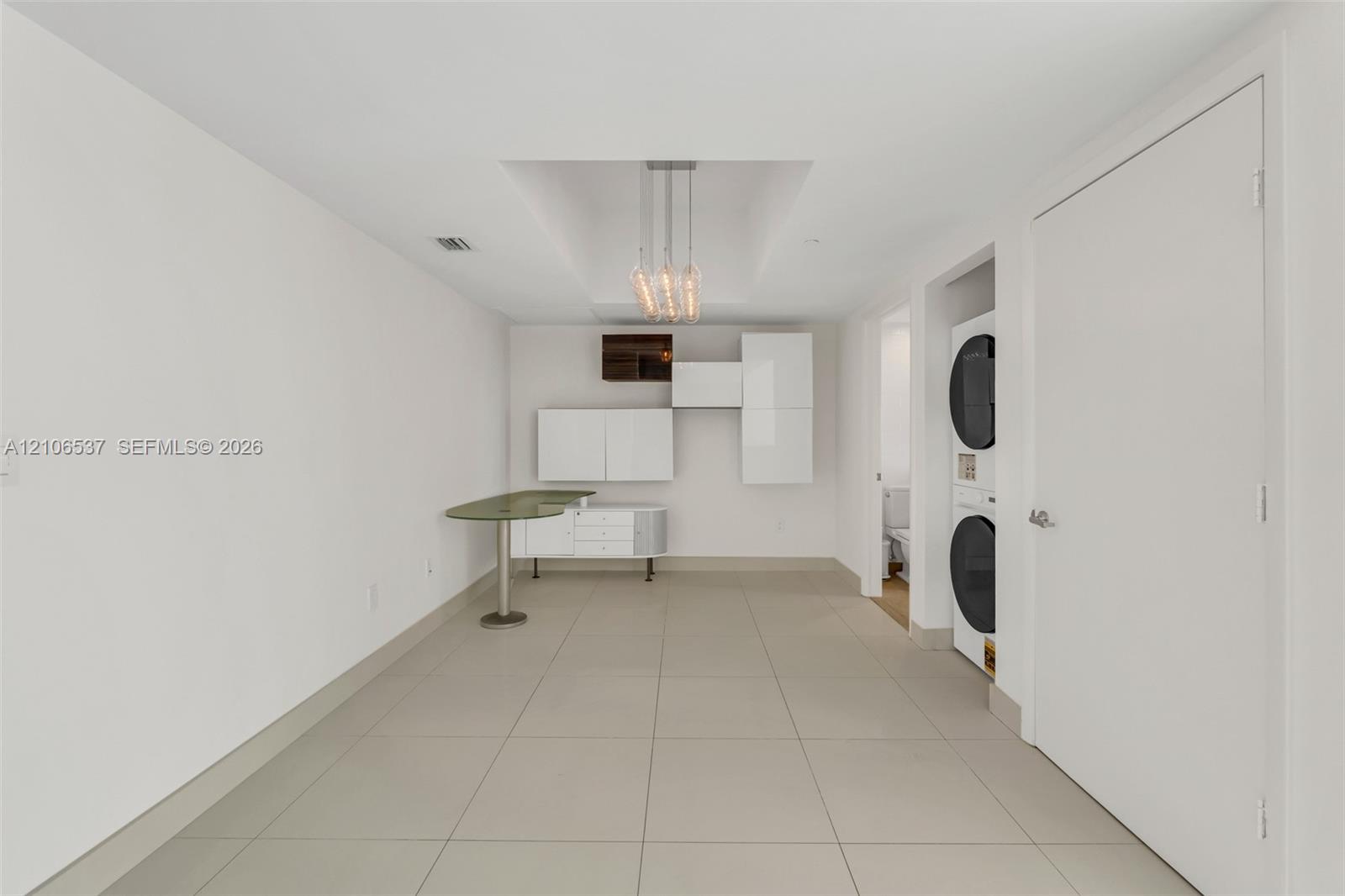 Modern utility space featuring large-format tile flooring, built-in glossy white cabinetry, an integrated stone-surface workstation, a tiered pendant light fixture, and a stacked washer and dryer unit