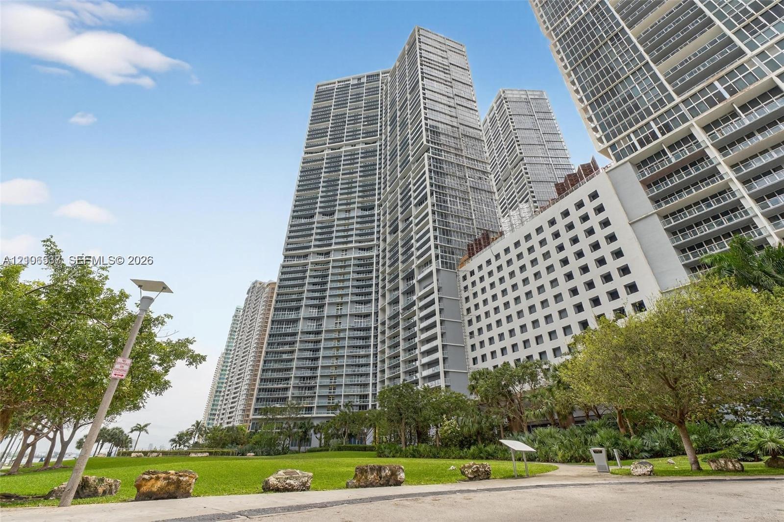 High-rise glass and concrete tower featuring tiered balconies, floor-to-ceiling windows, an expansive manicured lawn, and lush mature trees