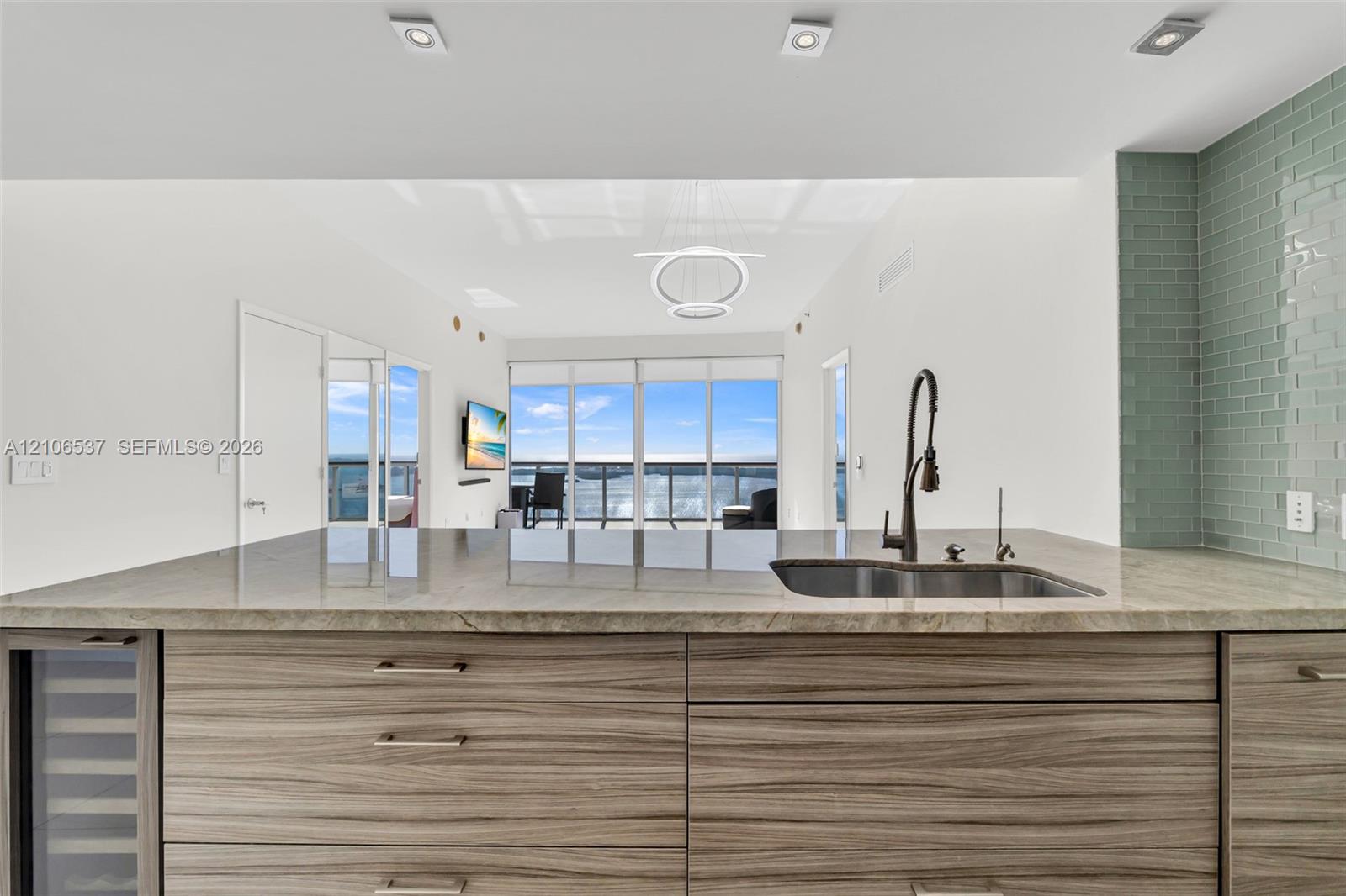 Kitchen island featuring wood-finish cabinetry, stone countertop, undermount sink with a matte black pull-down faucet, and a built-in wine cooler, complemented by a light green subway tile backsplash