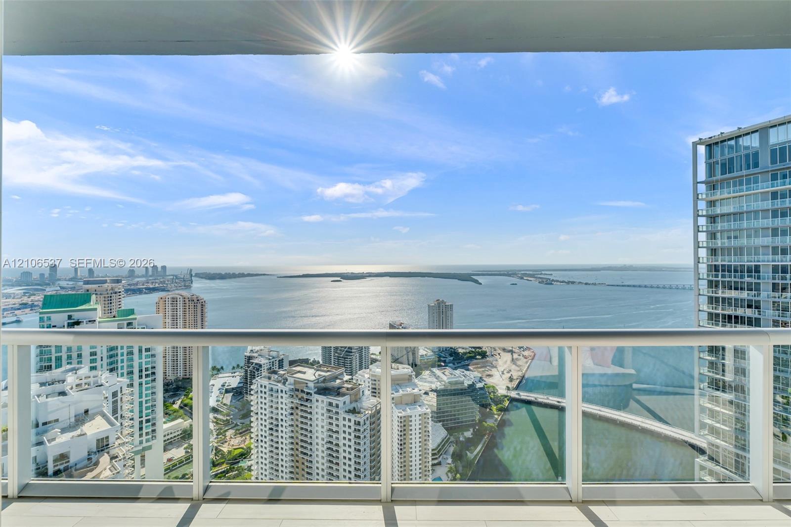 Covered balcony with a glass-panel railing, tile flooring, and expansive water and urban skyline backdrop Covered balcony with a glass-panel railing, tile flooring, and expansive water and urban skyline backdrop