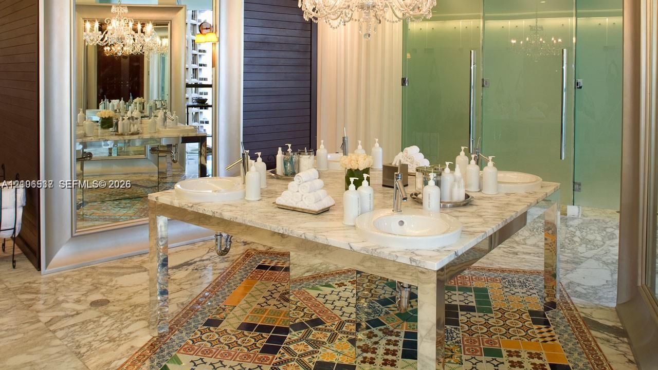 Custom vanity featuring a natural stone countertop, dual undermount sinks, and polished metal legs, complemented by decorative geometric tile flooring and a large-scale crystal chandelier