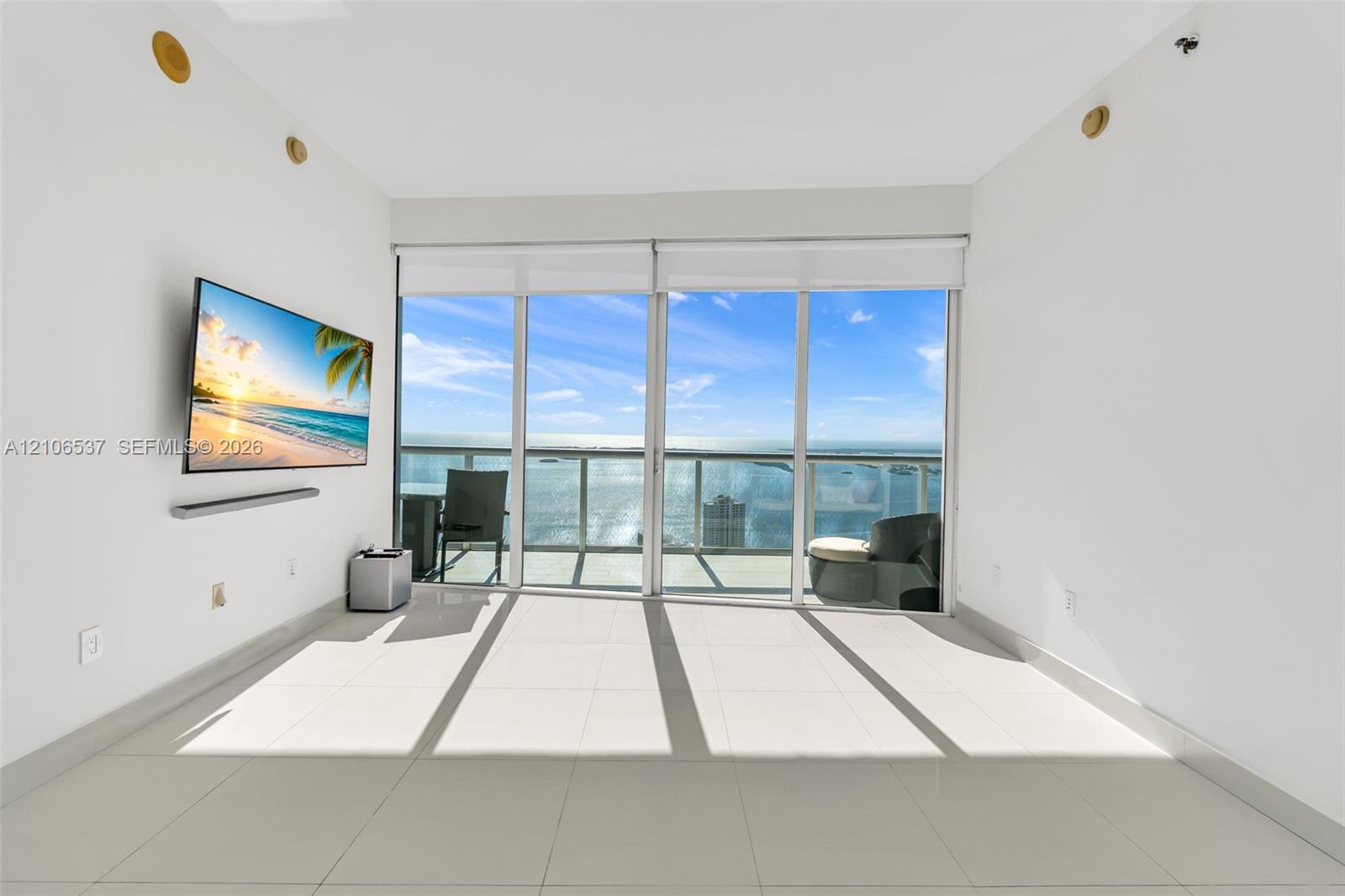 Floor-to-ceiling glass sliding doors, large-format tile flooring, and an attached balcony with metal railing, set against a waterfront backdrop