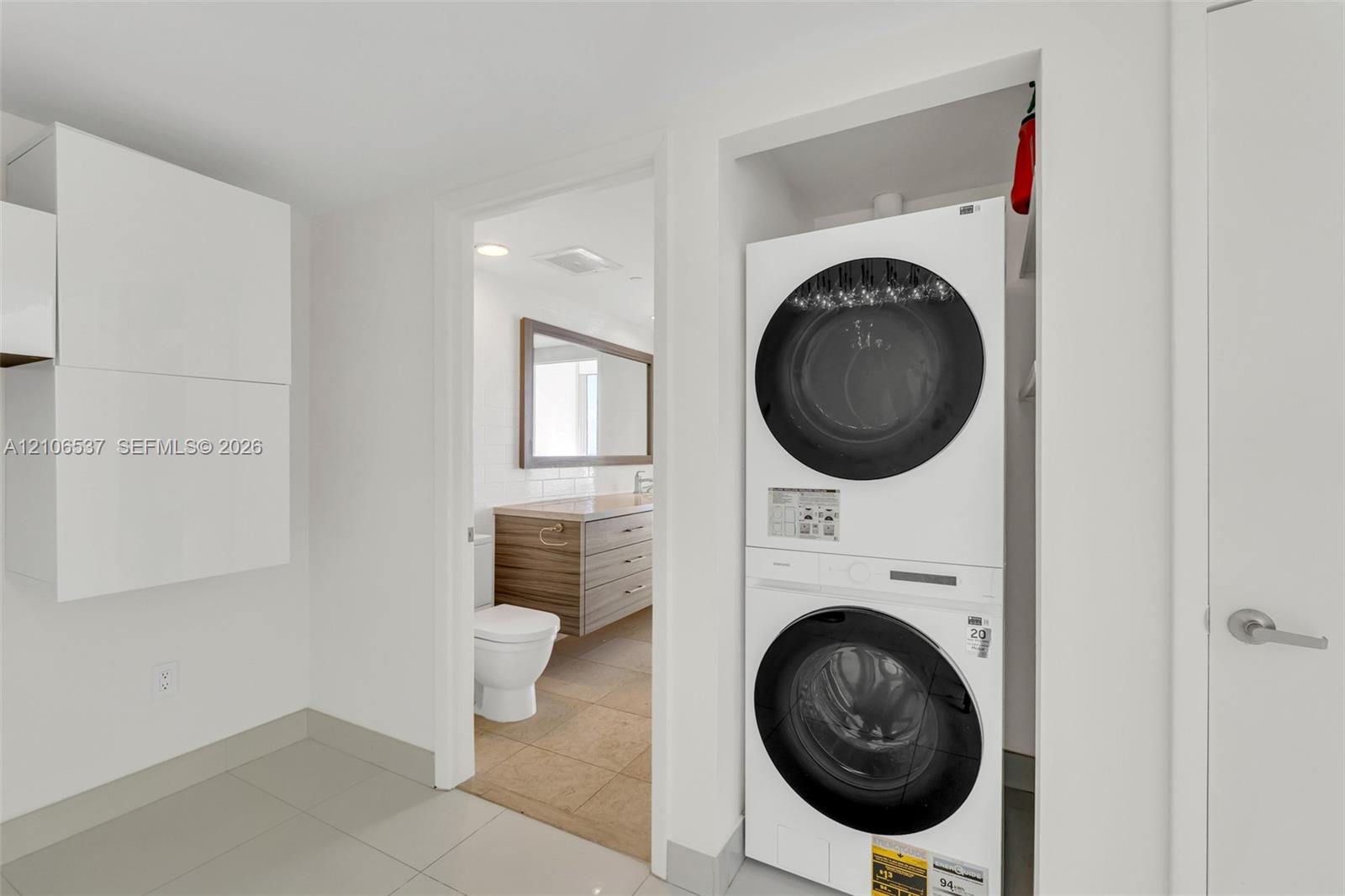 Laundry area featuring a stacked washer and dryer unit, built-in cabinetry, ceramic tile flooring, and a doorway to a bathroom equipped with a wood-finish vanity and rectangular mirror