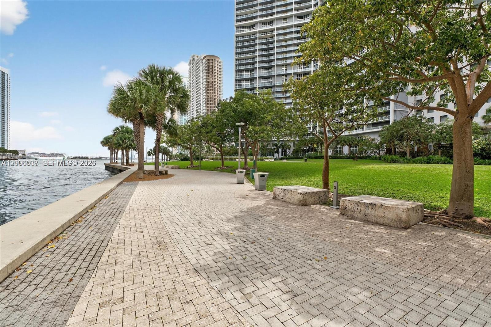 Waterfront paved walkway featuring stone seating, mature trees, manicured lawn areas, and concrete curbing
