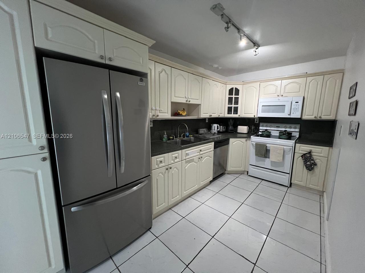 Kitchen featuring off-white cabinetry, stainless steel French-door refrigerator, dark solid-surface countertops, white tile flooring, and track lighting