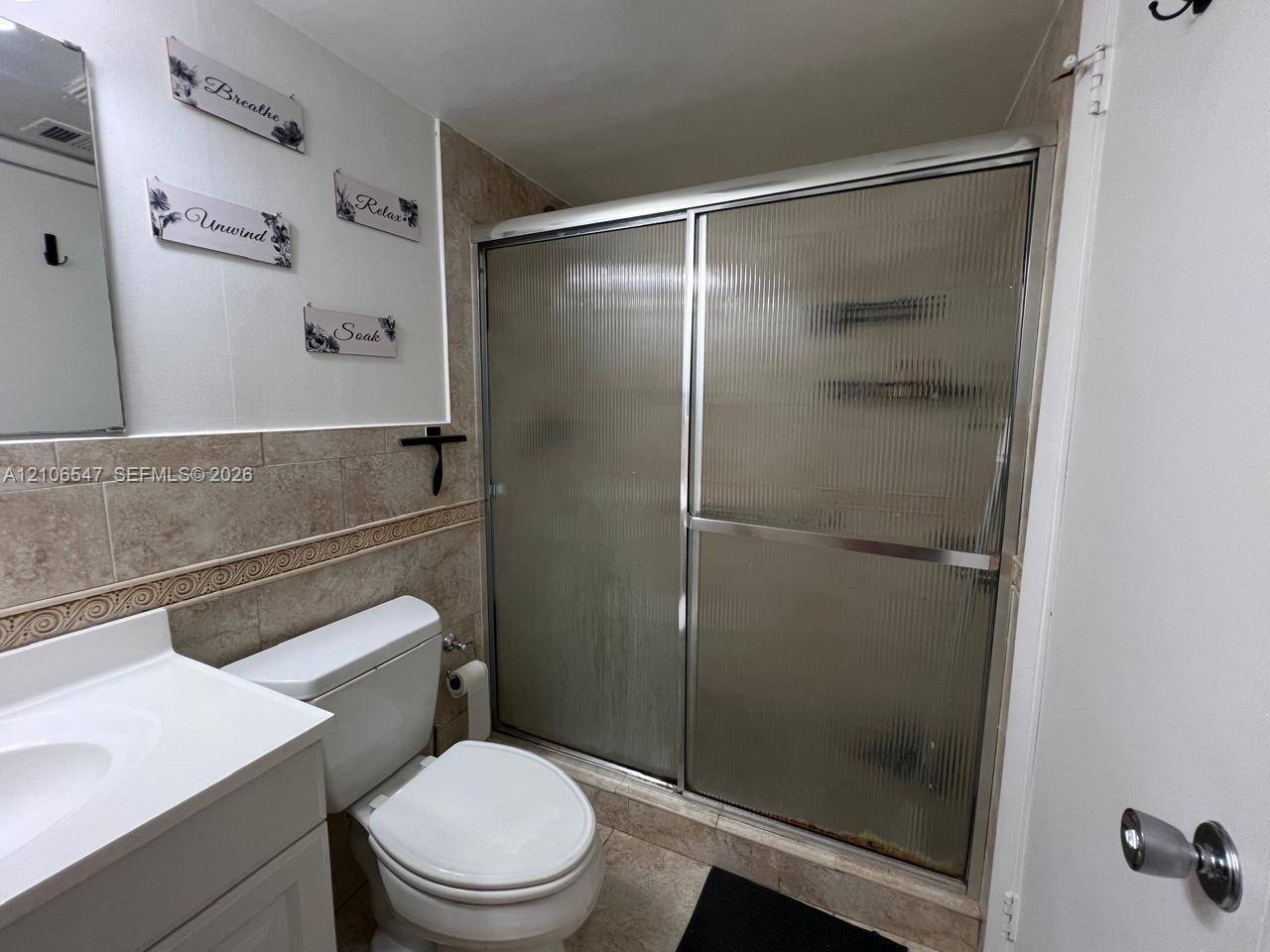 Bathroom featuring a white vanity with an integrated basin, a standard toilet, and a walk-in shower enclosed by reeded glass sliding doors with metallic framing, finished with wall-to-wall ceramic tile surround and coordinating floor tile