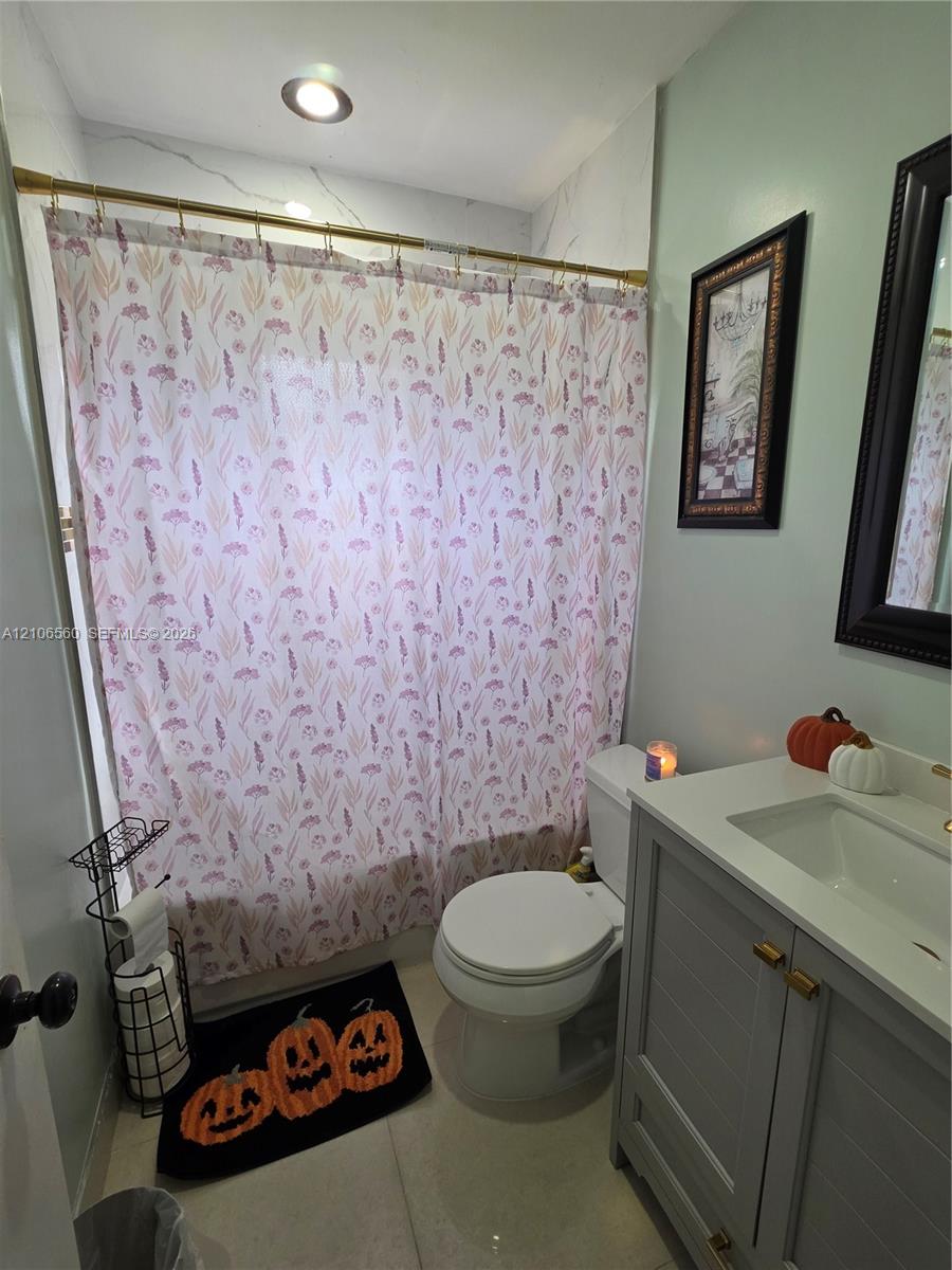 Grey shaker-style vanity with a solid surface countertop, integrated sink, brass hardware, and a tiled shower surround featuring a brass rod