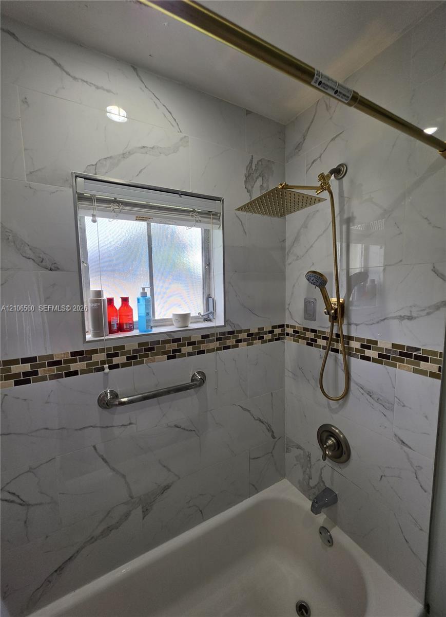 Bathtub surround featuring floor-to-ceiling stone-pattern tile, a multi-tone mosaic accent border, a gold-tone rainfall showerhead with a handheld sprayer, and a mounted metal grab bar