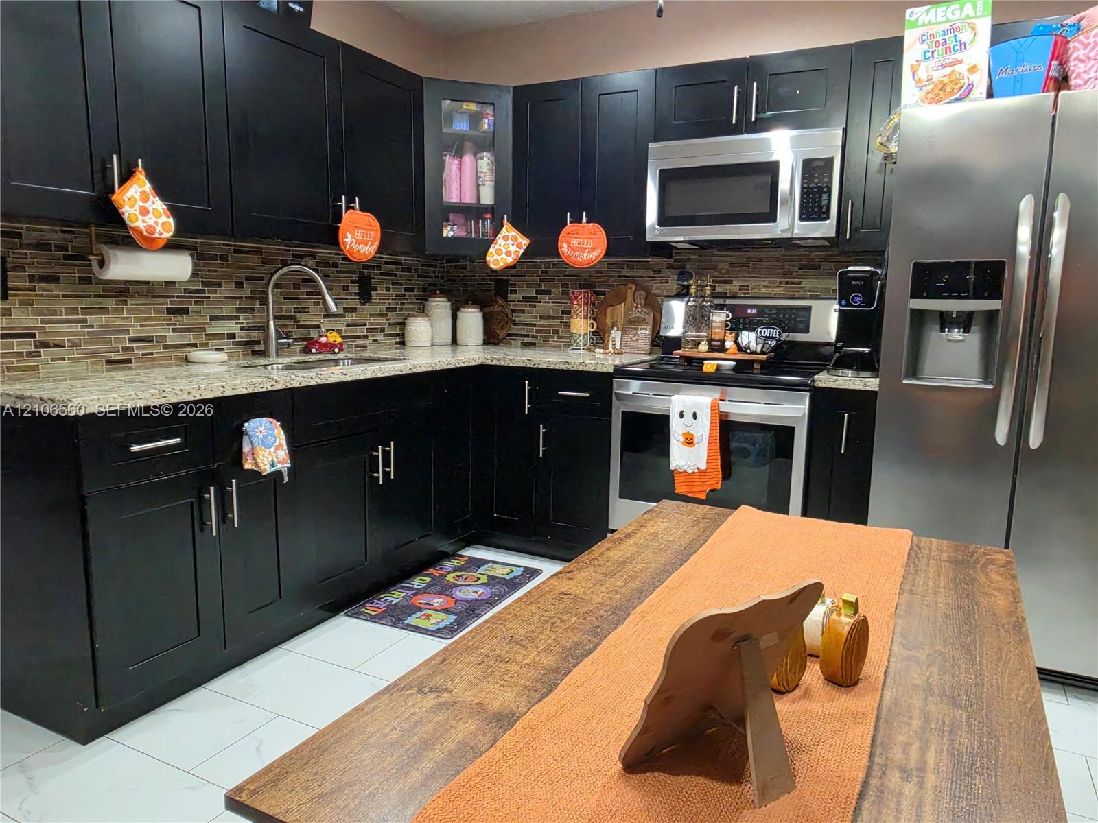 Kitchen featuring black cabinetry, light-toned stone-look countertops, a mosaic tile backsplash, stainless steel appliances, and tile flooring