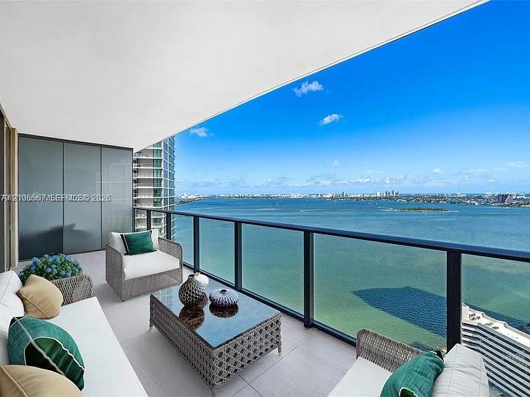 Covered balcony featuring neutral tile flooring, glass panel railings with dark metal framing, and extensive water exposure Covered balcony featuring neutral tile flooring, glass panel railings with dark metal framing, and extensive water exposure