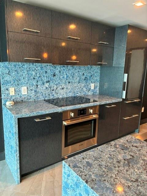 High-gloss dark cabinetry featuring brushed metal pulls, blue patterned stone countertops, matching full-height stone backsplash, and integrated stainless steel wall oven and cooktop