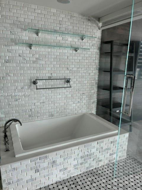 Built-in soaking tub with a tiled surround, complemented by a floor-to-ceiling accent wall in brick-pattern stone, dual glass wall-mounted shelves, and a wall-mounted metal towel rack