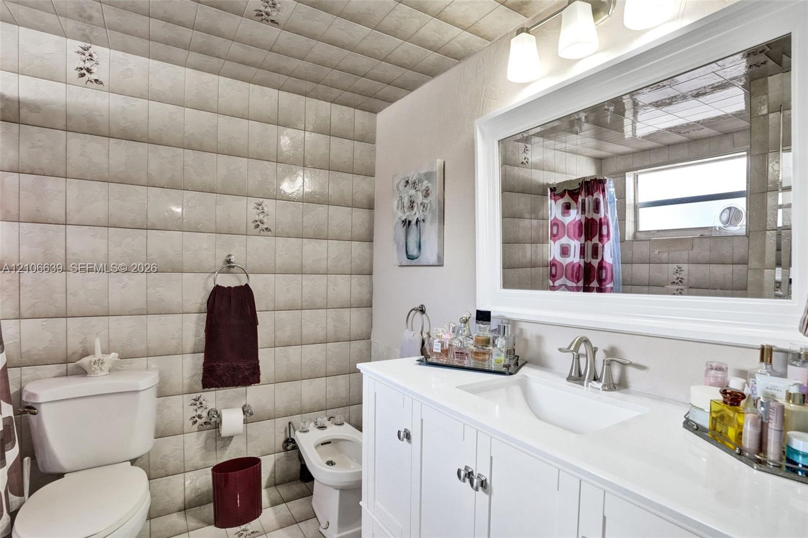 White vanity with a smooth integrated countertop and dual-handle faucet, expansive wall-mounted mirror, three-bulb vanity light fixture, bidet, and floor-to-ceiling decorative tiled wall finishes