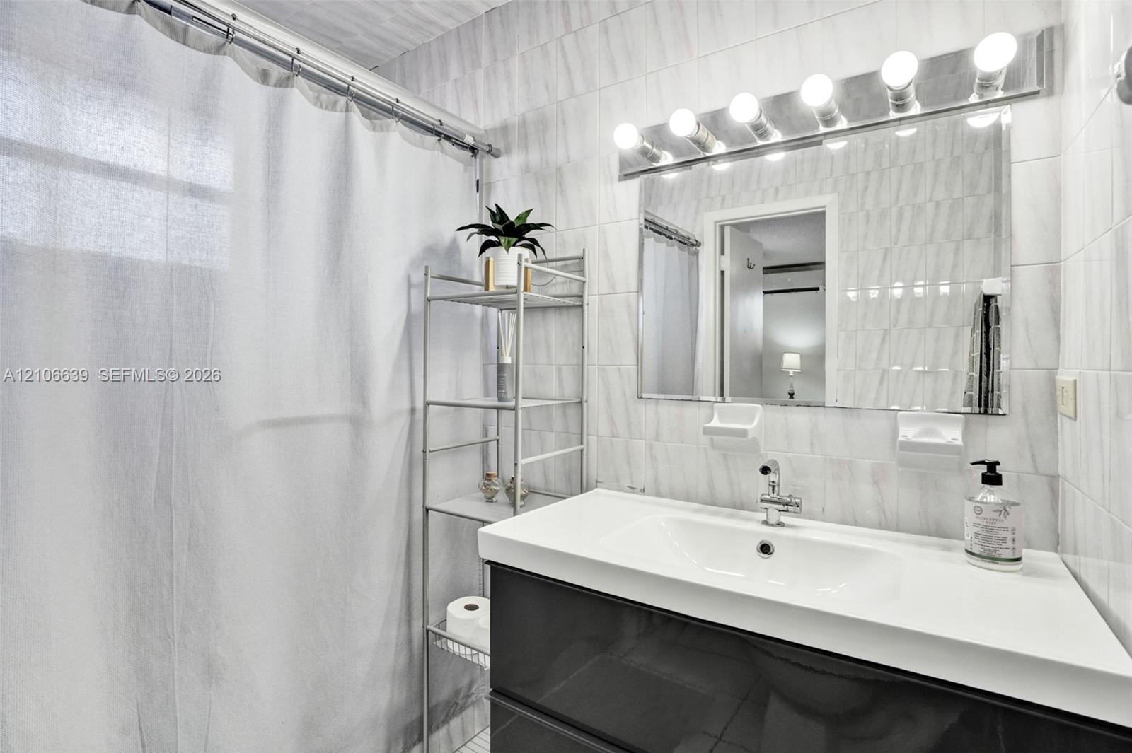 Wall-mounted vanity featuring a glossy dark cabinet, white integrated sink, chrome faucet, full-width mirror with six-bulb light fixture, and floor-to-ceiling wall tile