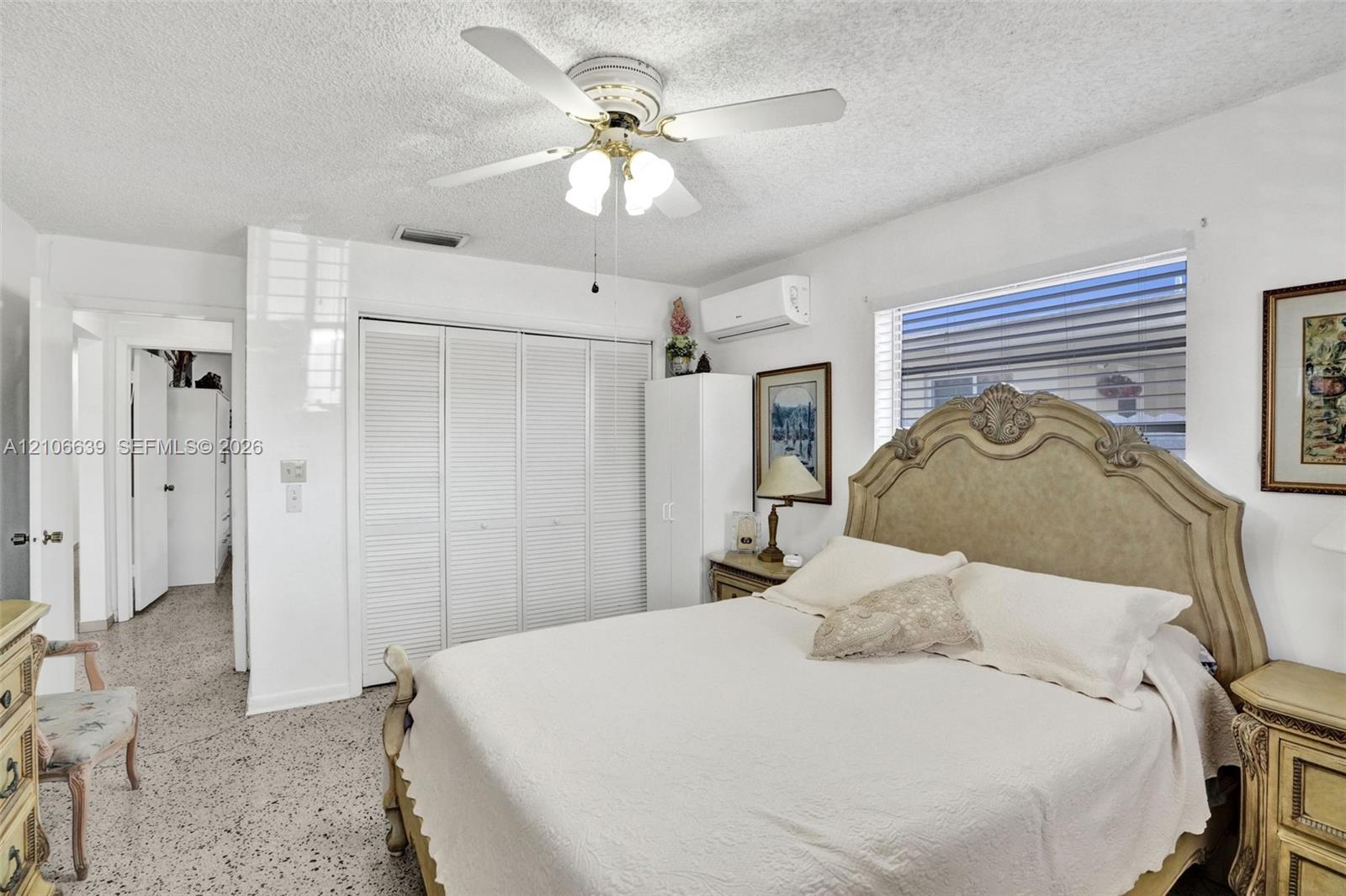 Multi-panel louvered closet doors, wall-mounted air conditioning unit, ceiling fan with integrated lighting, and speckled composition flooring