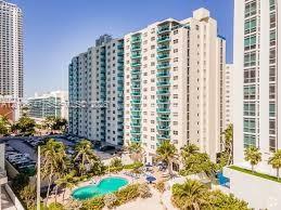 High-rise residential building featuring stacked private balconies with glass railings, an outdoor swimming pool, and landscaped grounds with mature palm trees