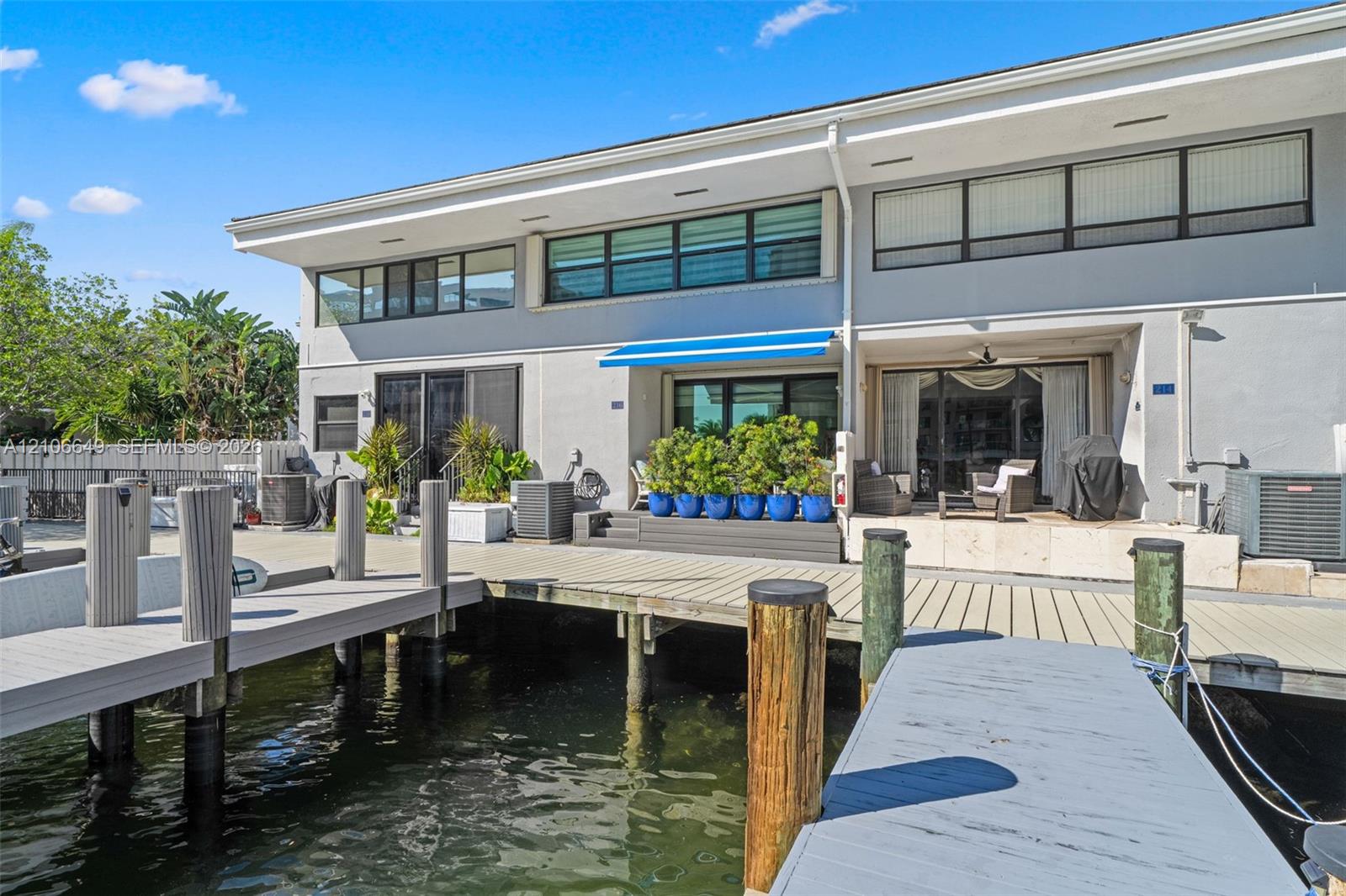 Waterfront exterior featuring a multi-level composite boat dock, stone-surfaced patio area, retractable blue awning, and large-format sliding glass doors
