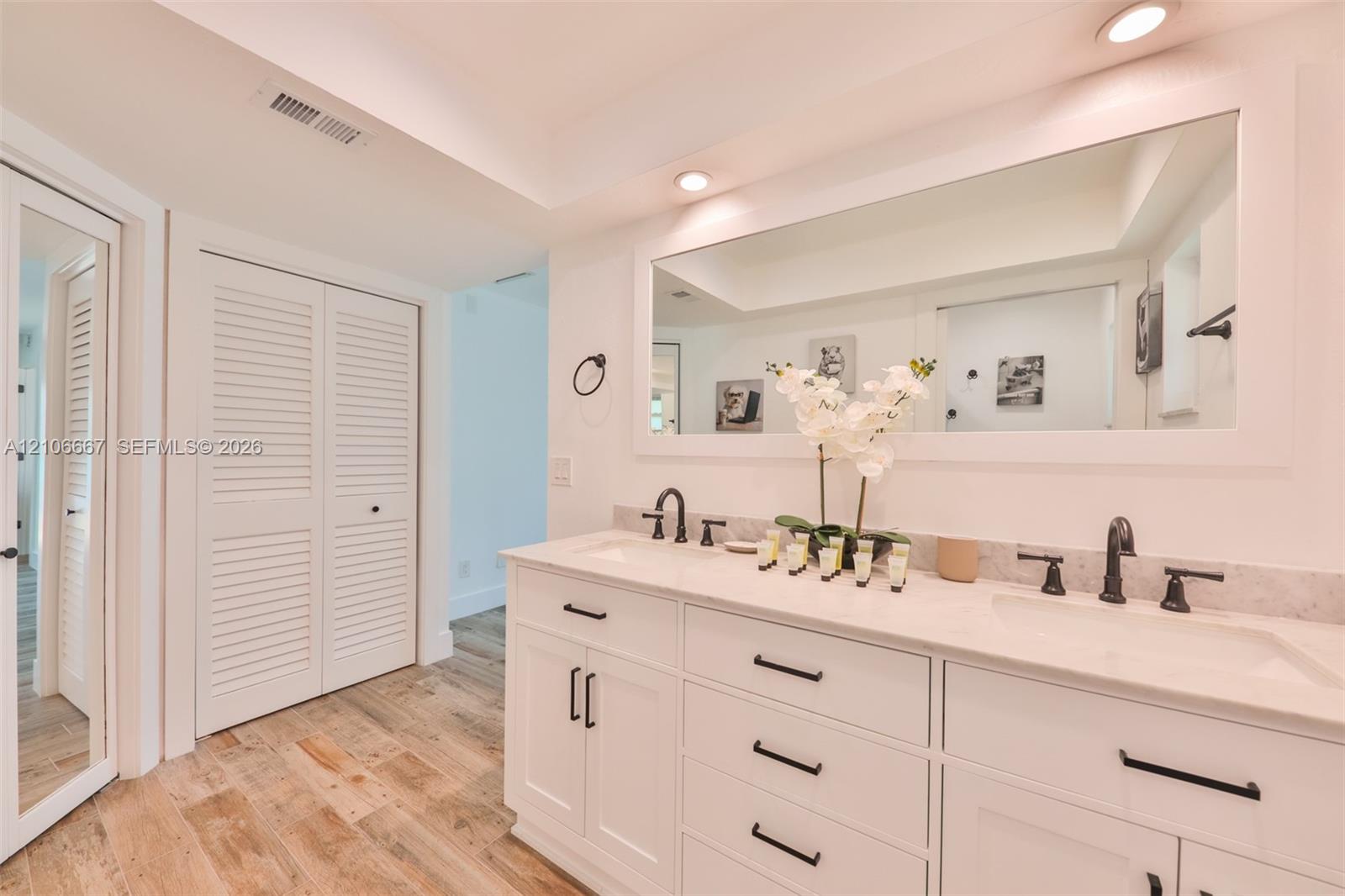 Dual-sink vanity featuring white cabinetry, stone-finish countertops, matte black hardware, and an oversized framed wall mirror, complemented by wood-finish flooring and louvered closet doors