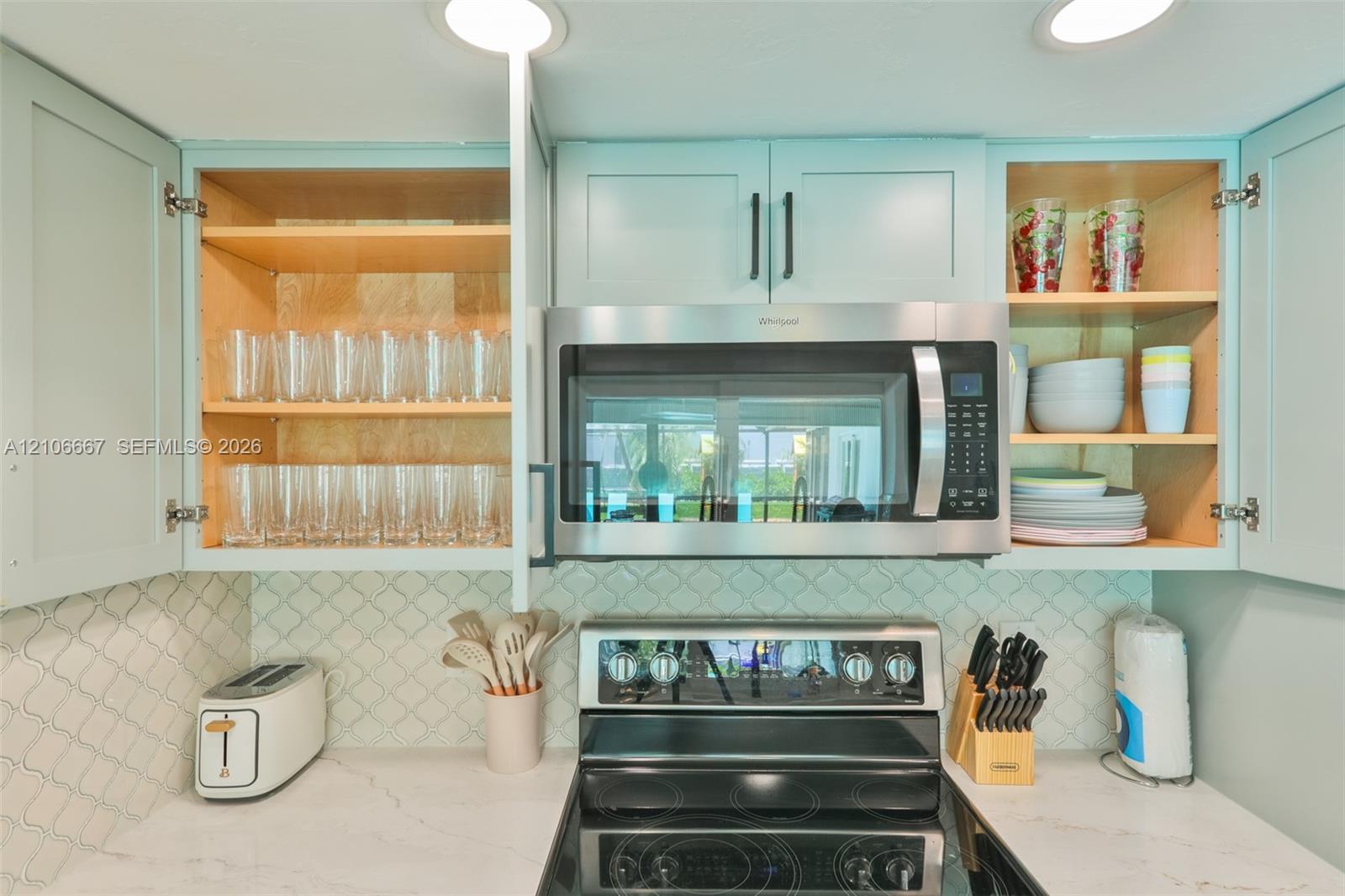 Custom cabinetry with light-toned finish, stainless steel over-the-range microwave, smooth-top electric range, patterned tile backsplash, and light-toned solid surface countertops with integrated recessed lighting