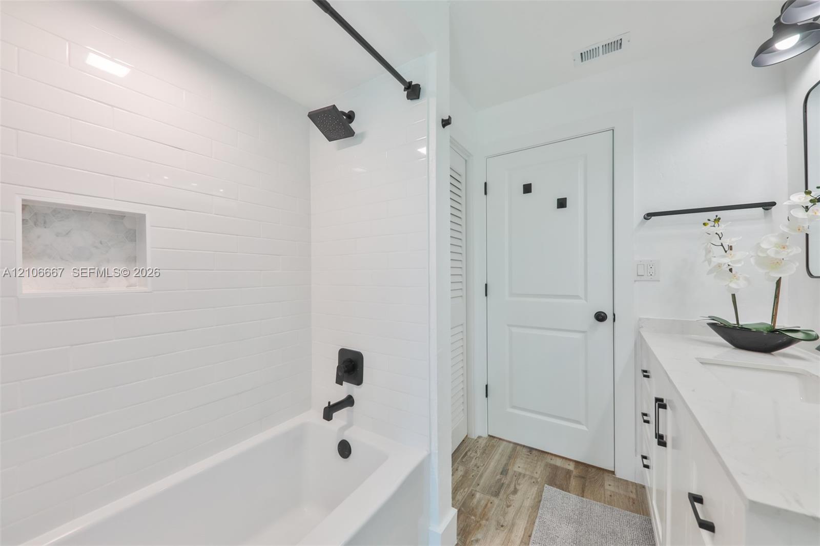 Bathroom featuring a white subway-tiled tub enclosure with an integrated recessed niche, matte black fixtures, white vanity with stone-finish countertop, and wood-finish flooring