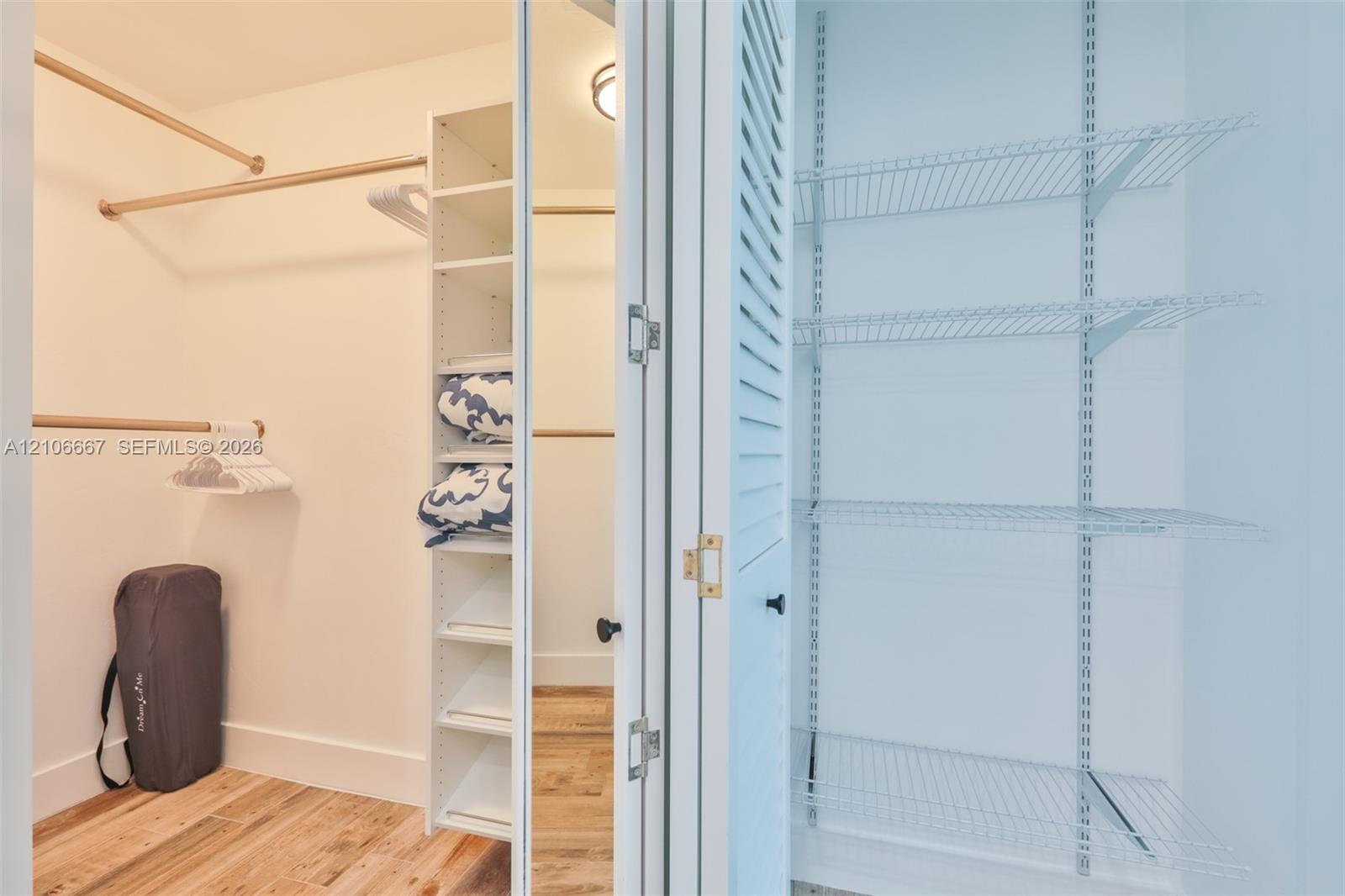 Walk-in closet featuring wood-finish flooring, built-in shelving tower, tiered hanging rods, and a separate pantry-style storage area with louvered doors and wire shelving
