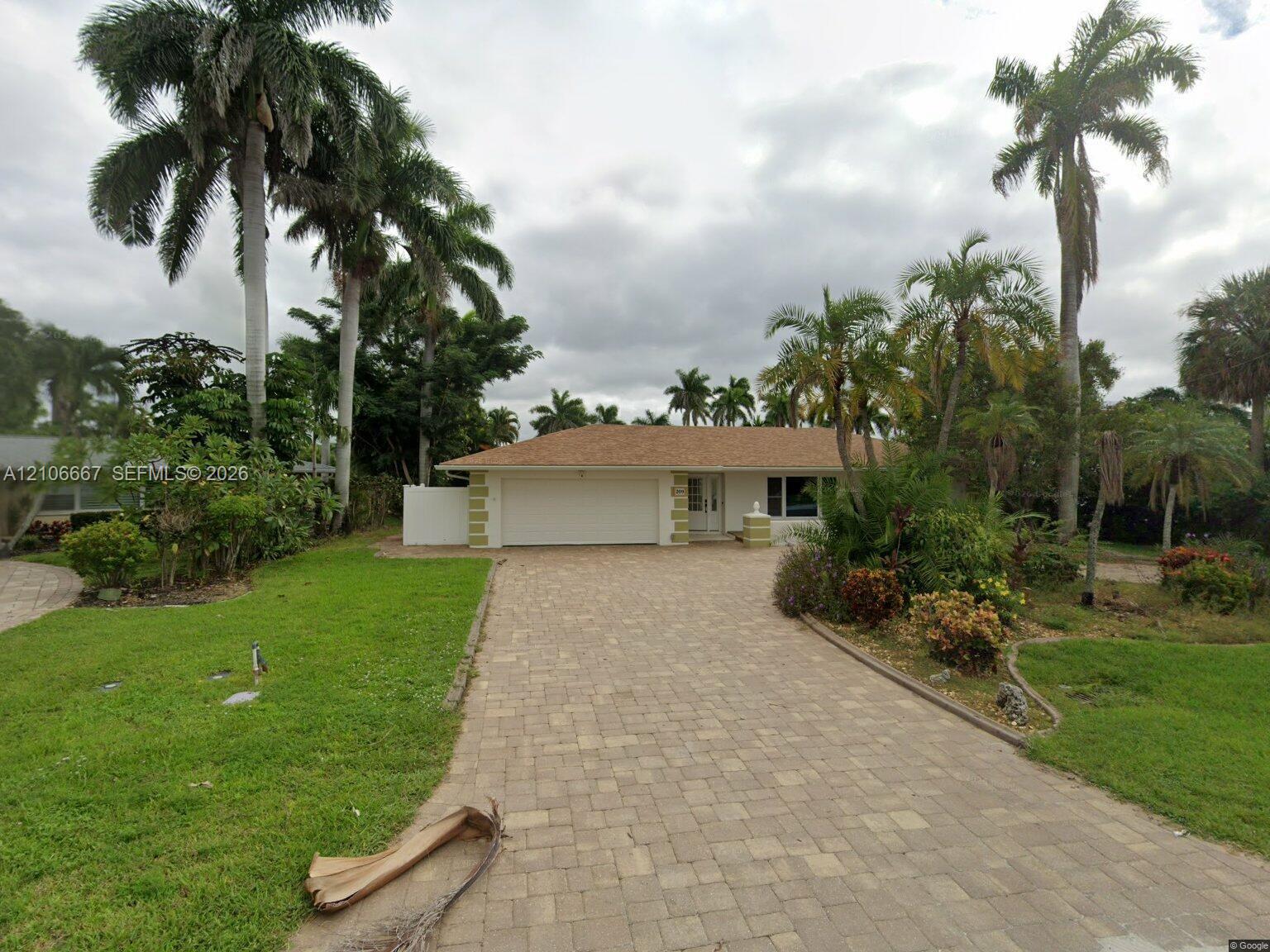 Single-story residence featuring a shingle roof, paved stone driveway, attached two-car garage, and established landscaping with mature palm trees Single-story residence featuring a shingle roof, paved stone driveway, attached two-car garage, and established landscaping with mature palm trees