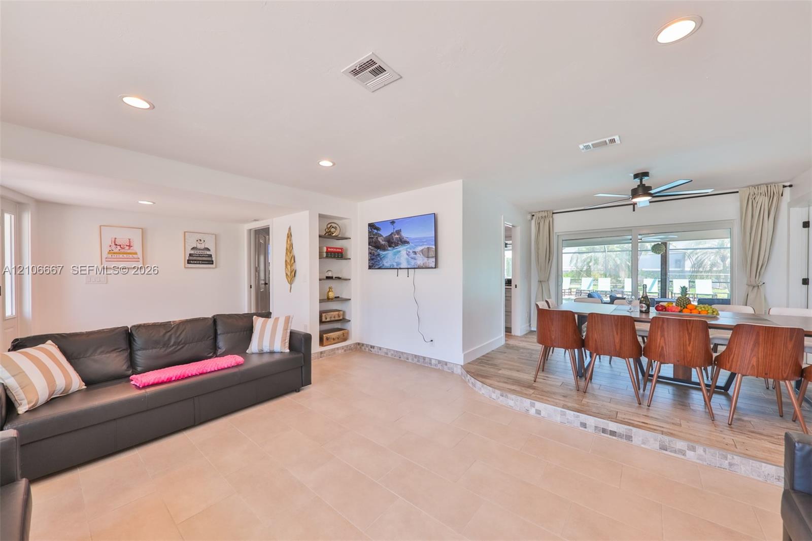 Open-concept interior featuring light-toned tile flooring, recessed lighting, built-in wall shelving, wood-finish raised platform, and large sliding glass doors Open-concept interior featuring light-toned tile flooring, recessed lighting, built-in wall shelving, wood-finish raised platform, and large sliding glass doors