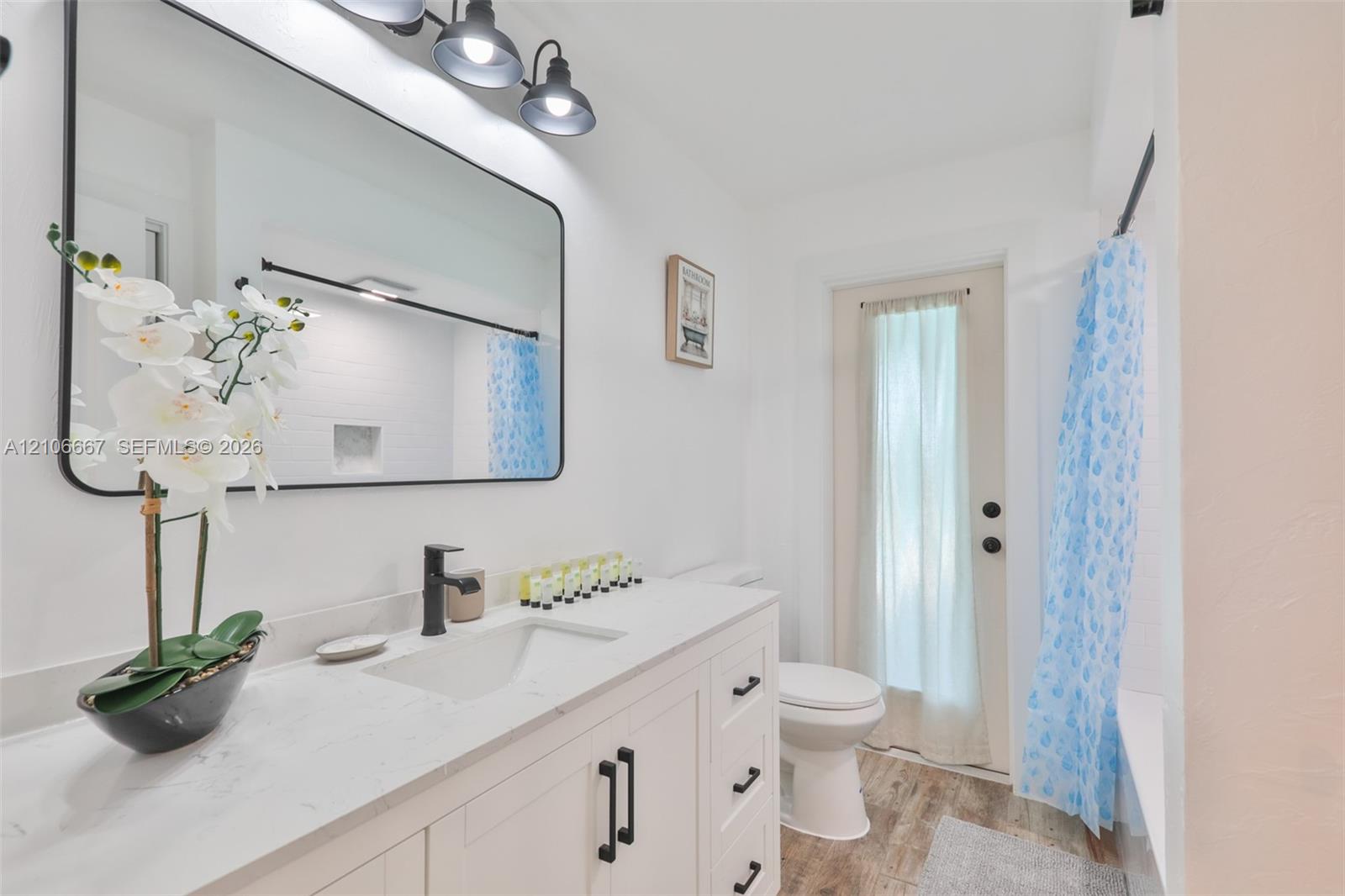 Bathroom featuring a vanity with a white countertop and rectangular integrated sink, matte black faucet and hardware, rectangular mirror with rounded corners, three-light vanity fixture, and wood-finish flooring