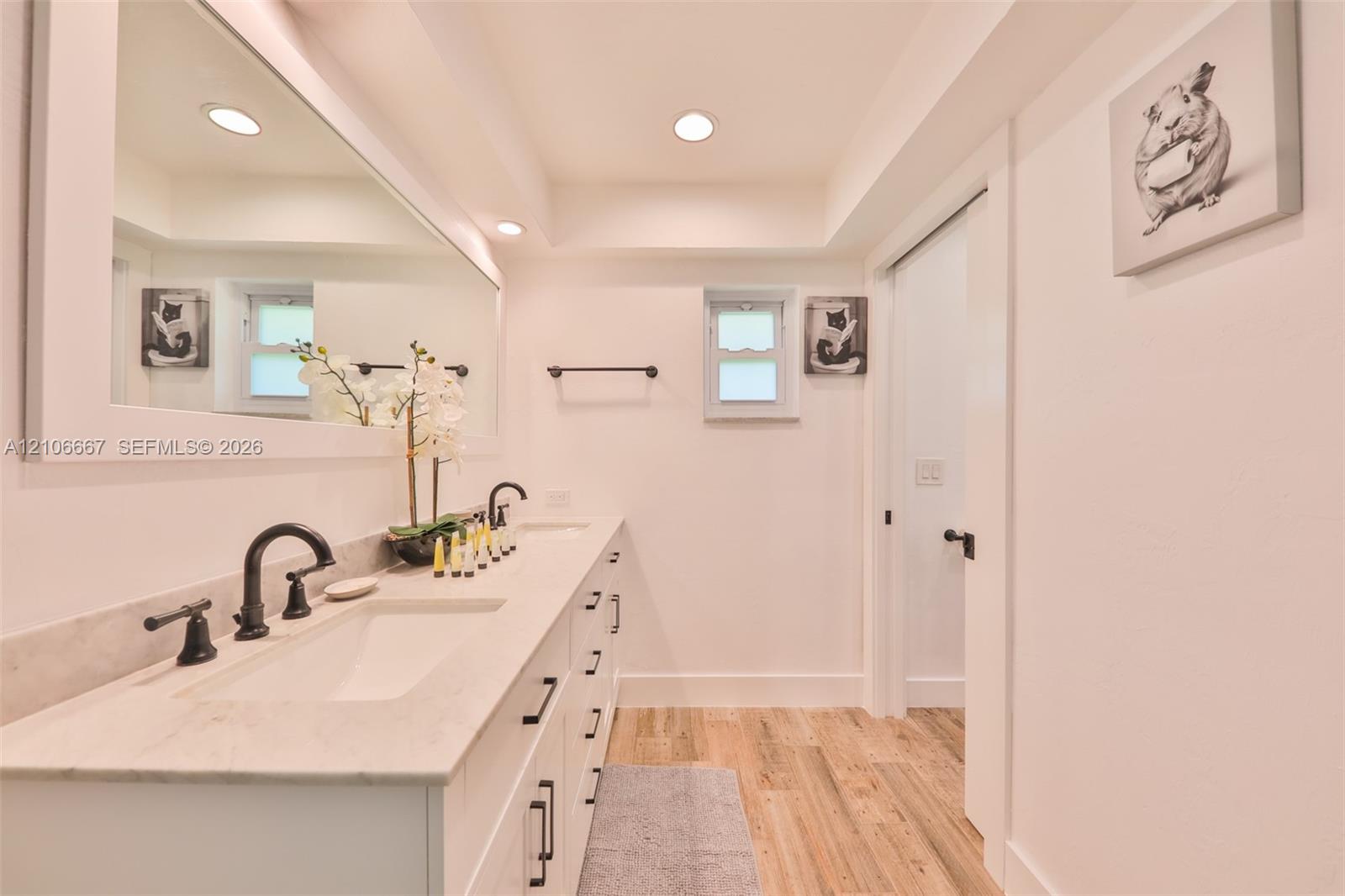 Double vanity featuring a light-toned countertop with dual integrated sinks, matte black widespread faucets, full-width framed mirror, and wood-finish flooring