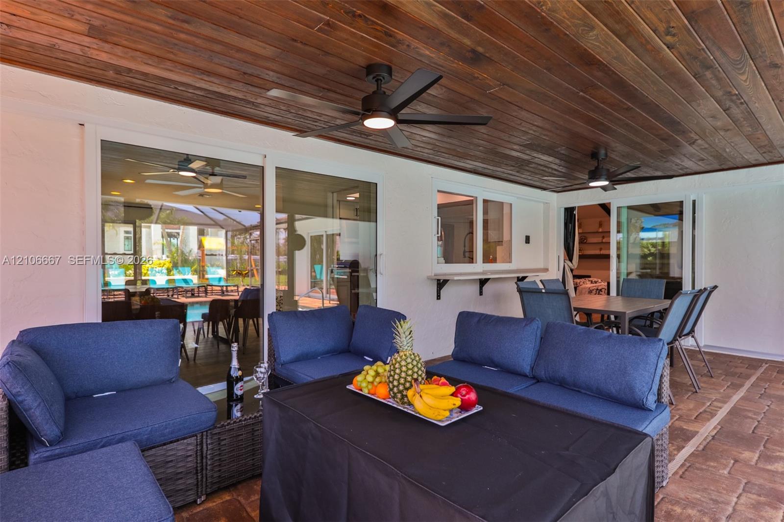 Covered patio featuring a wood-plank ceiling, matte black ceiling fans, paver flooring, sliding glass wall panels, and a built-in exterior pass-through window