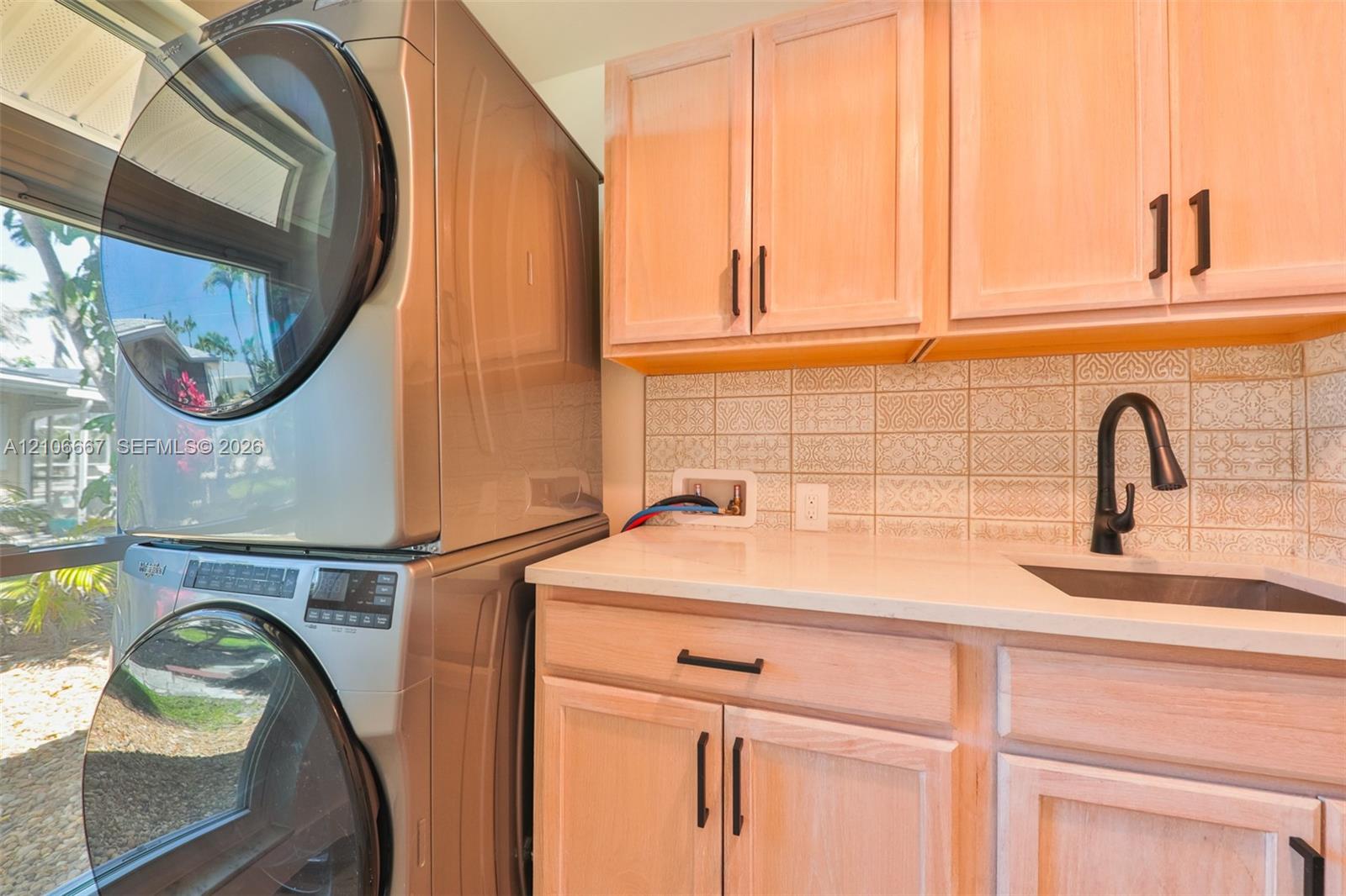 Laundry area featuring light-finish wood cabinetry, matte black hardware, stone-composite countertops, patterned tile backsplash, and a drop-in sink with a high-arc faucet