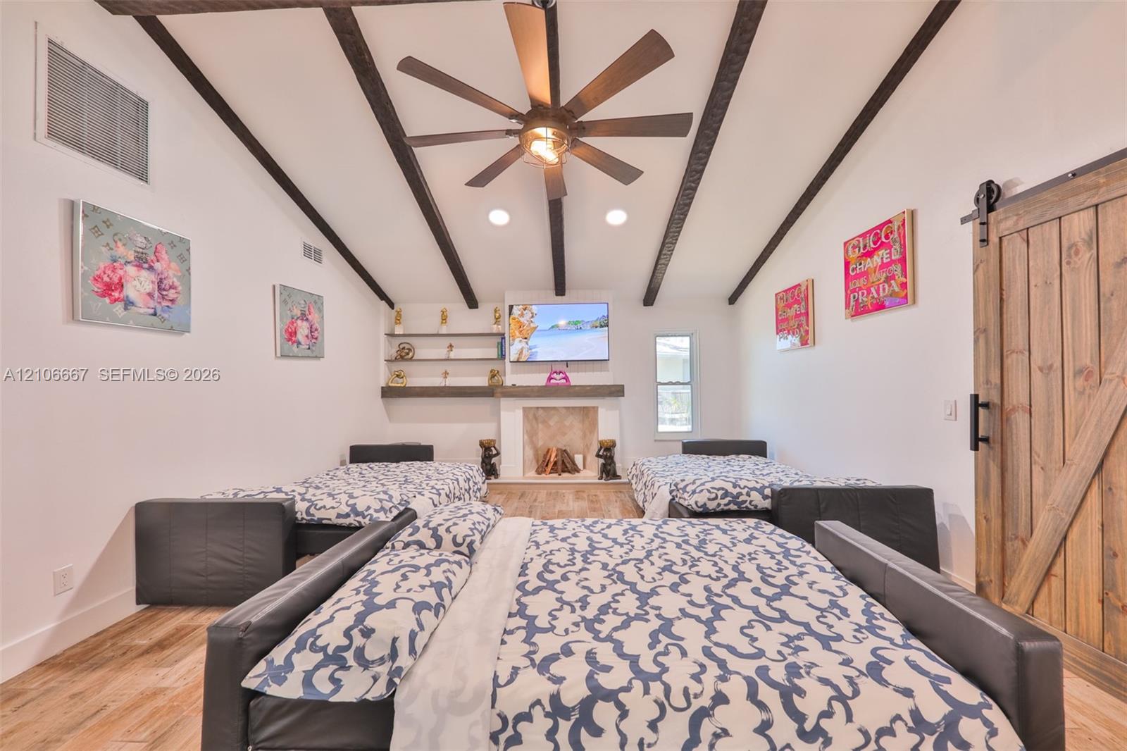 Vaulted ceiling with exposed decorative beams, wood-finish flooring, fireplace with stone-surround and mantle, built-in shelving, and sliding barn door