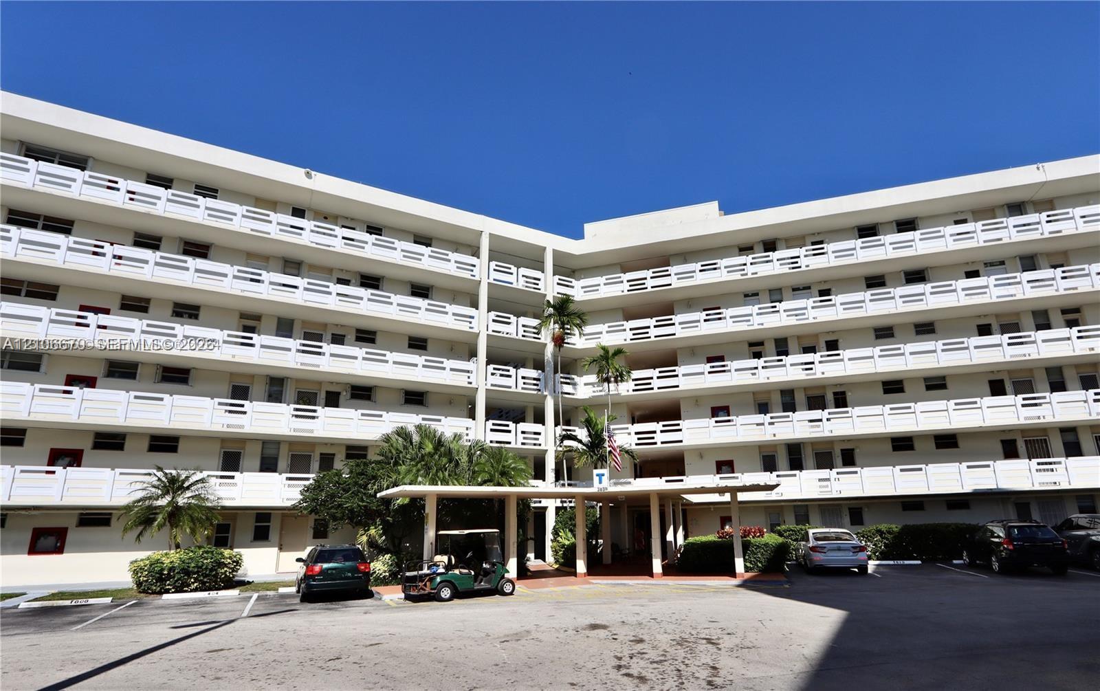 Multi-story building exterior featuring white concrete balconies with geometric railings, a covered porte-cochre entry, and mature palm tree landscaping