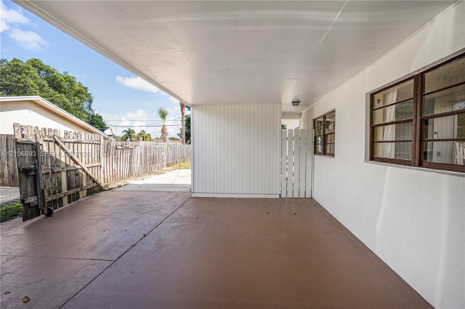 Covered concrete carport featuring white vertical siding, single-hung windows with metal frames, and a white privacy gate Covered concrete carport featuring white vertical siding, single-hung windows with metal frames, and a white privacy gate