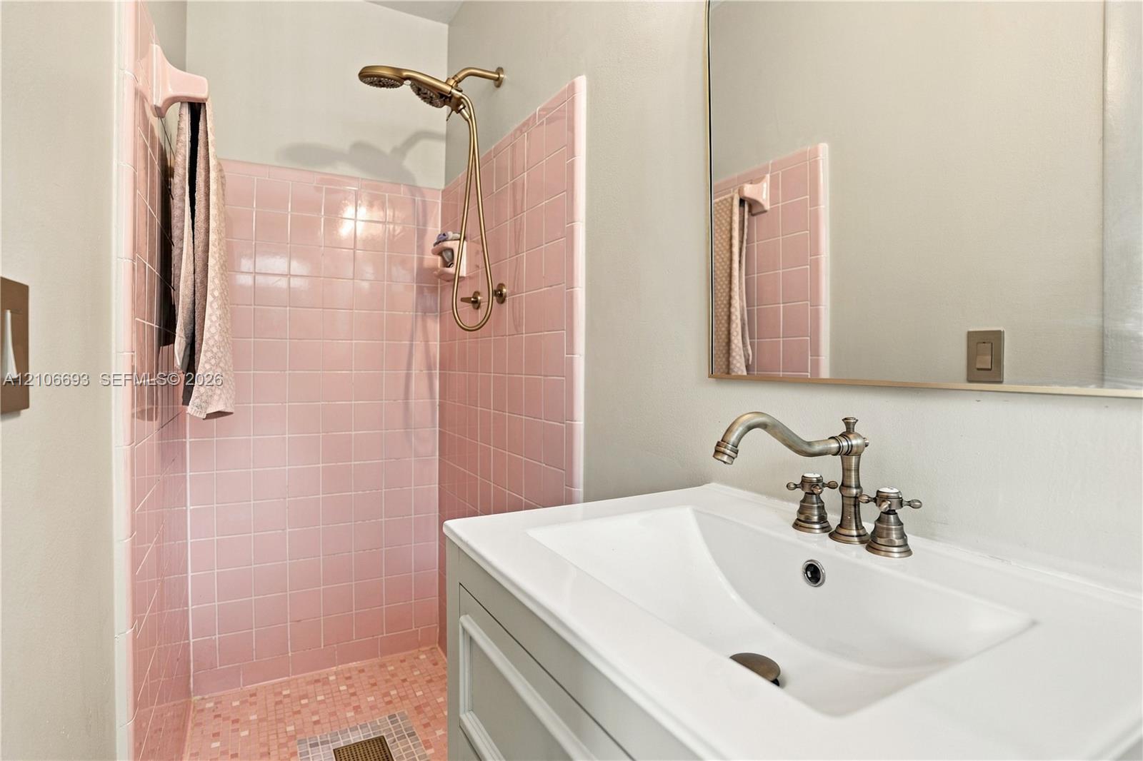 Bathroom featuring square pink tile shower surround, brass-finish handheld shower fixture, rectangular white undermount sink, brass-finish widespread faucet, and large frameless mirror Bathroom featuring square pink tile shower surround, brass-finish handheld shower fixture, rectangular white undermount sink, brass-finish widespread faucet, and large frameless mirror
