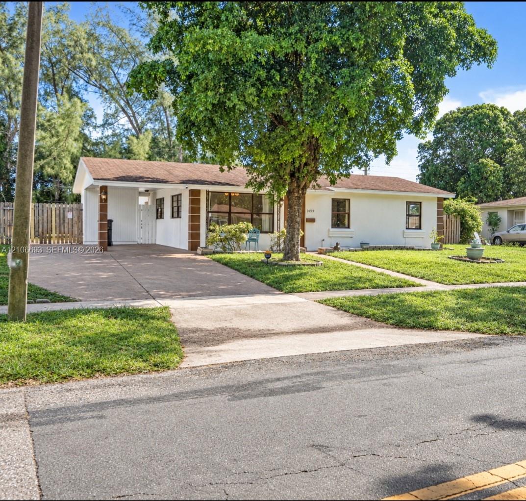 Single-story exterior featuring light-toned stucco siding, brown masonry accents, an attached covered carport, a concrete driveway, and mature landscaping